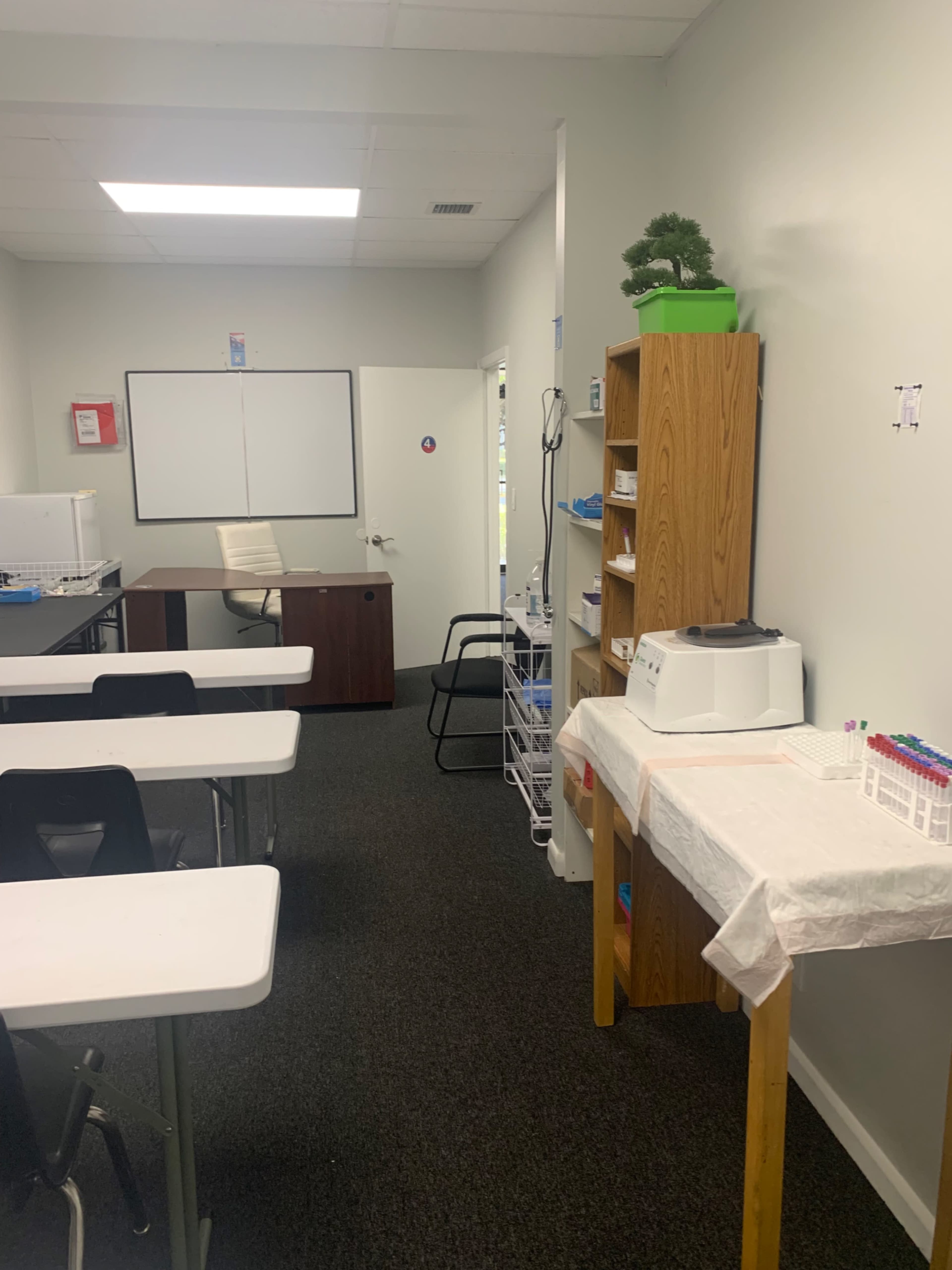 A classroom with several white tables and chairs, a wooden storage unit, and a desk at the back against a light-colored wall.