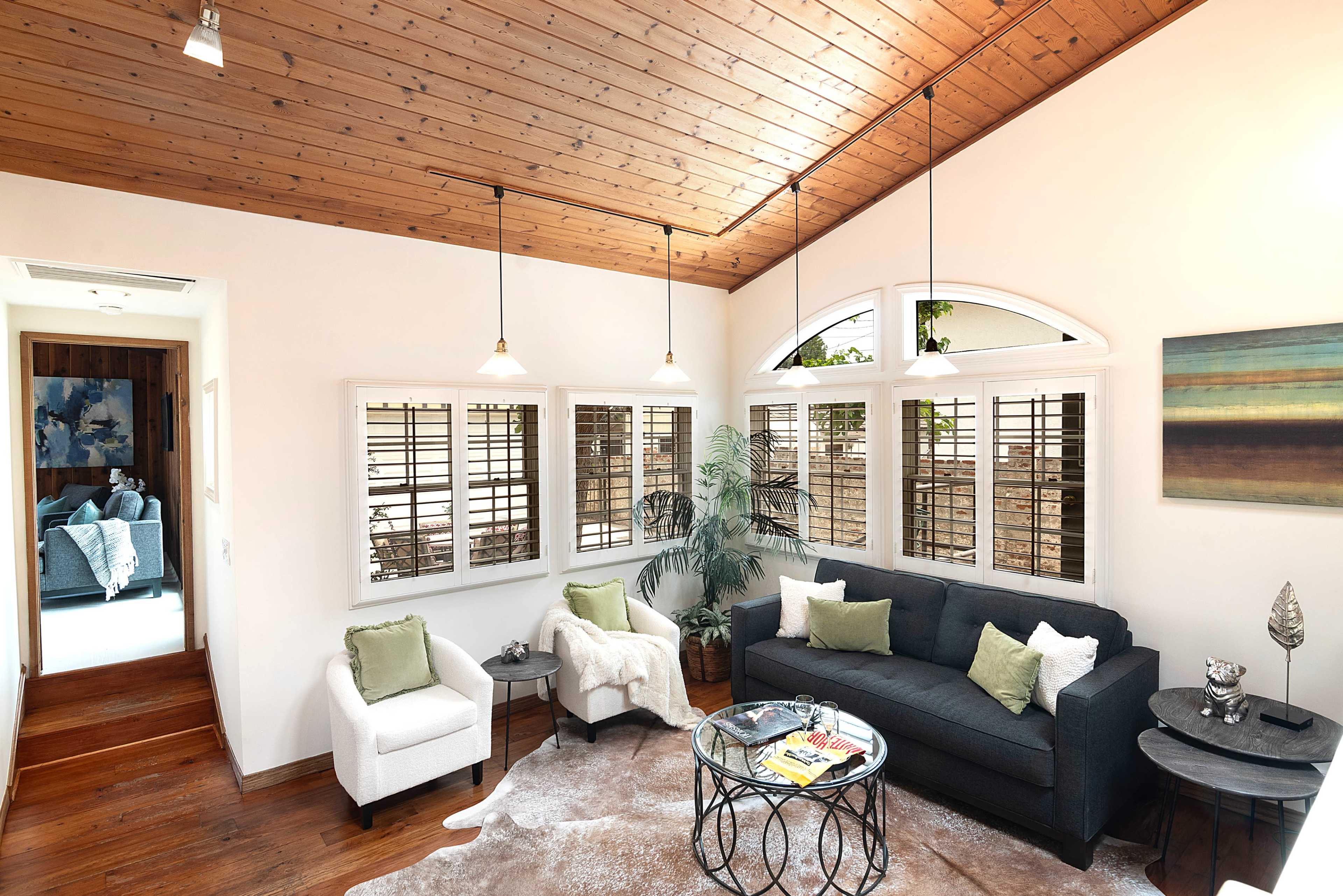 A living room features a dark sofa, two white chairs, and a round coffee table, with large windows allowing natural light and wooden accents on the ceiling.