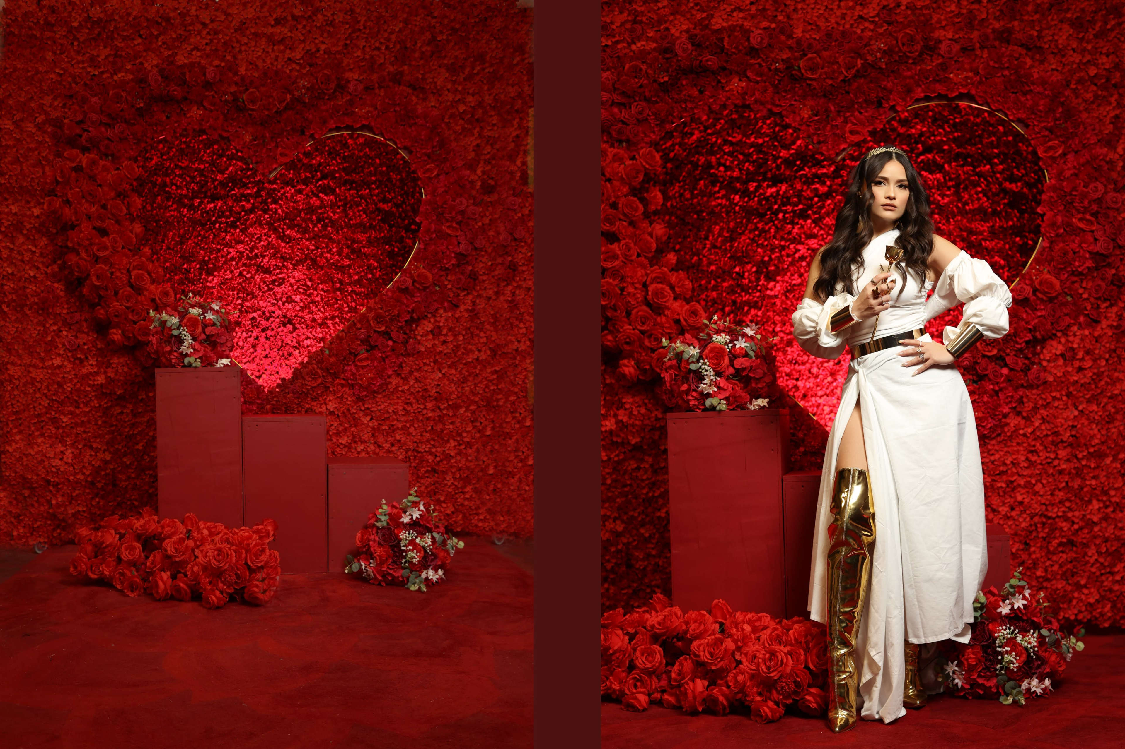 A woman in a white outfit with a thigh-high slit stands beside a red heart-shaped floral backdrop, surrounded by decorative boxes and red roses.
