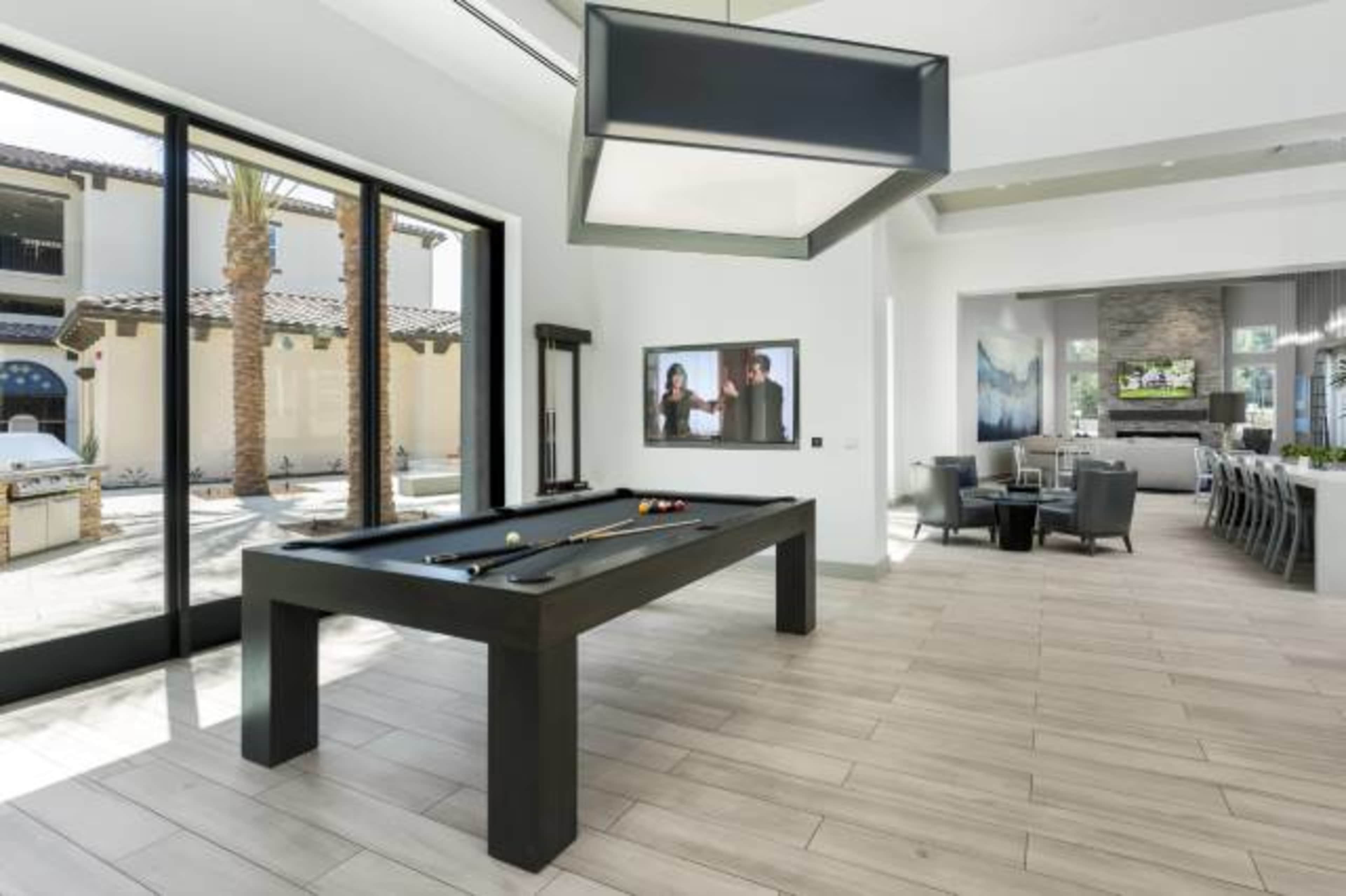 A game room features a black pool table and large windows overlooking a courtyard with palm trees.