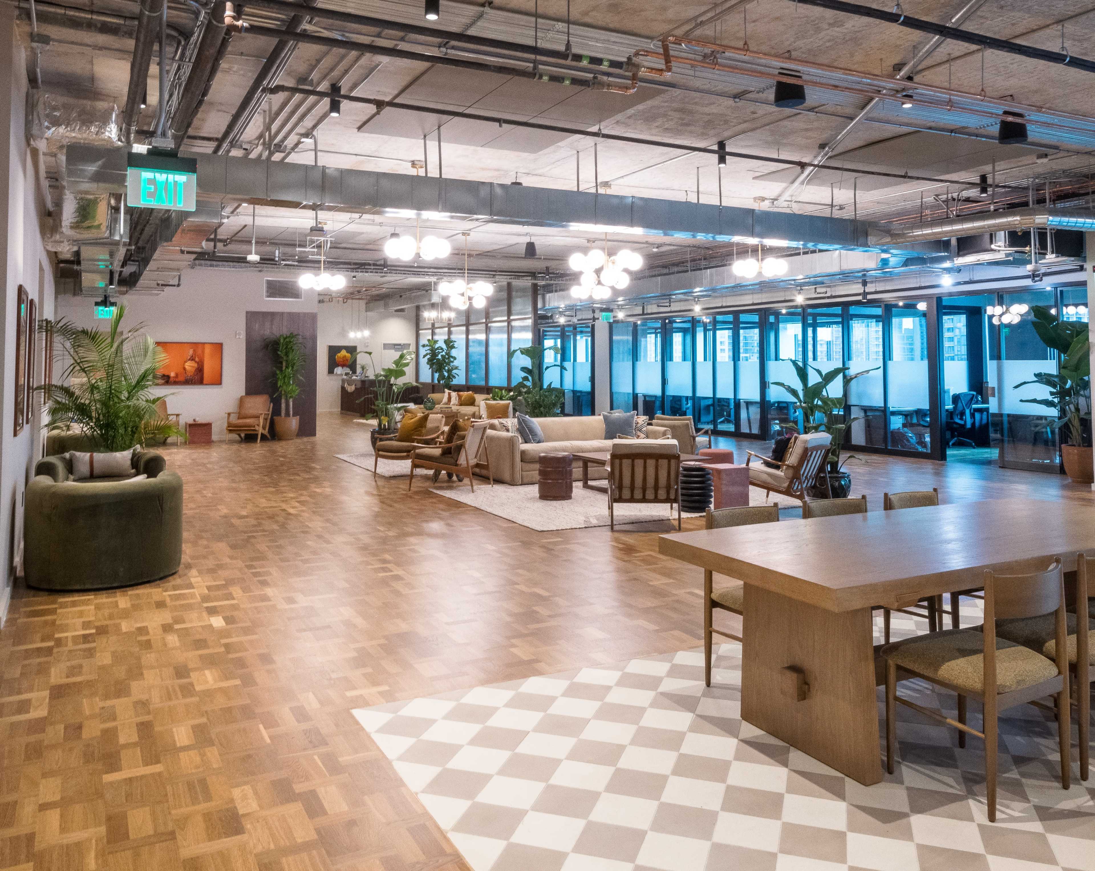 A modern office interior with various seating areas, a dining table, and large windows, featuring wooden floors and contemporary lighting.