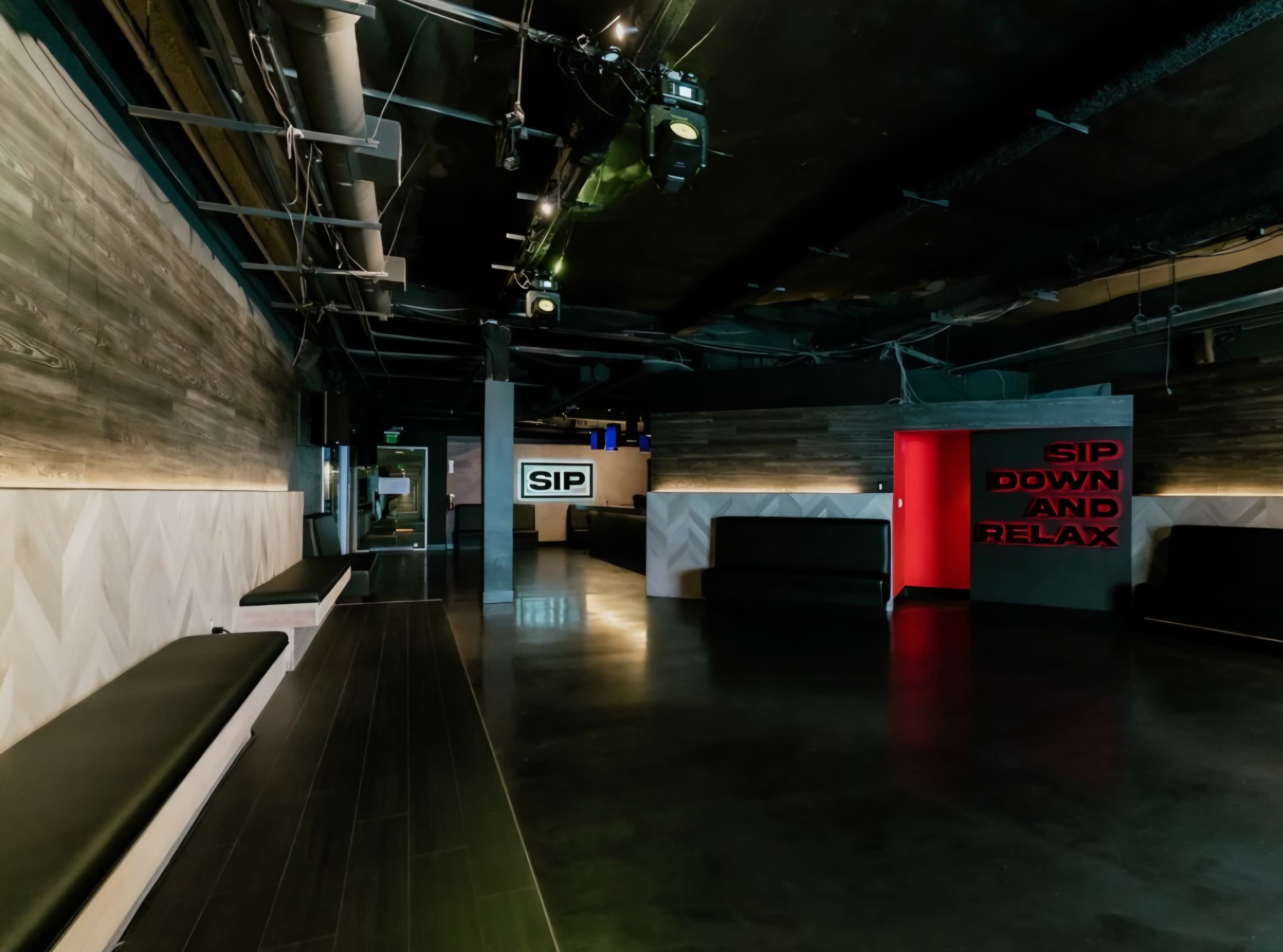 The image shows a modern, spacious interior of a venue with dark flooring, wooden walls, and benches along the sides, featuring illuminated signage that reads "SIP" and "SIP DOWN AND RELAX."