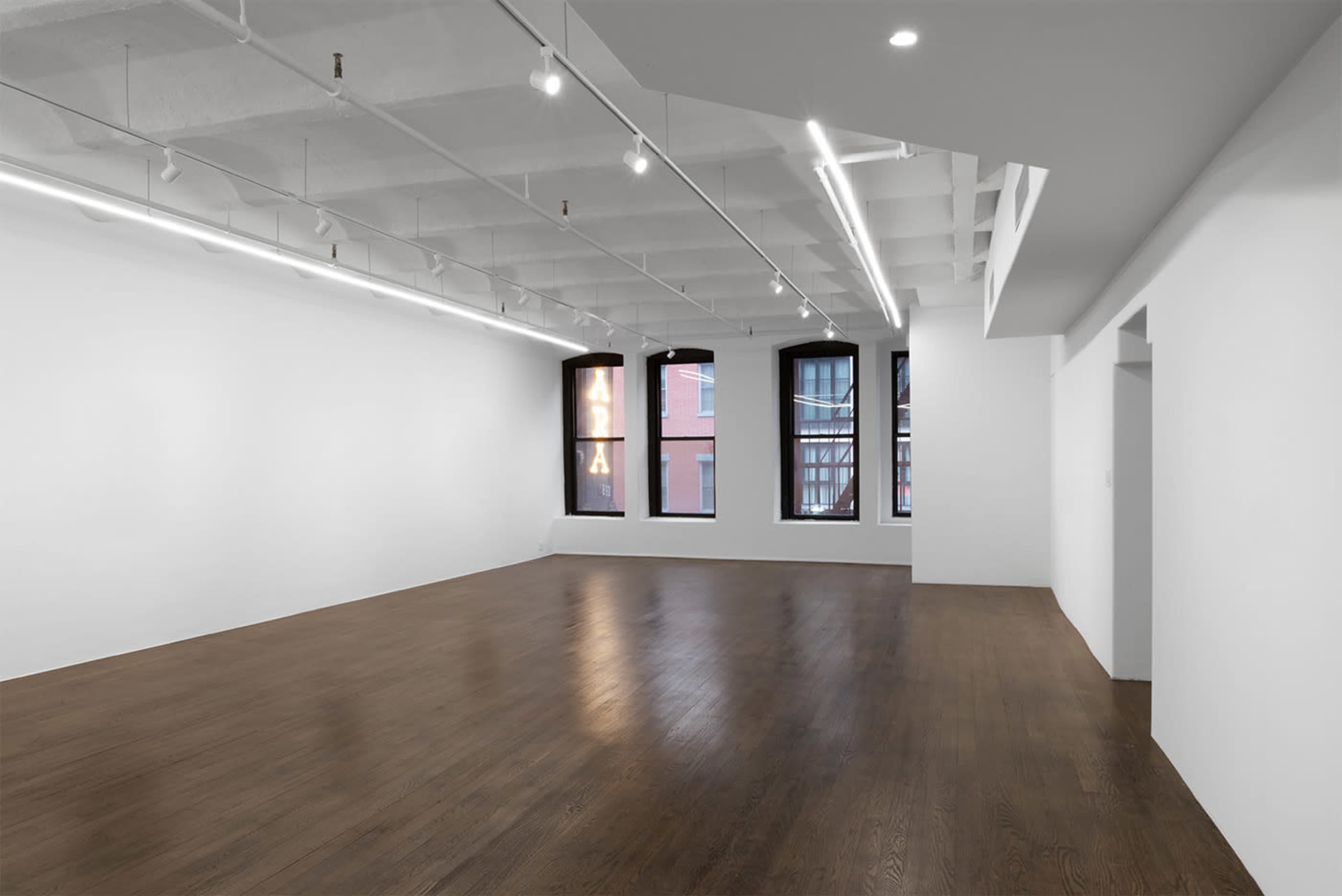 The image shows a large, empty gallery space with polished wooden floors and large windows along one wall.