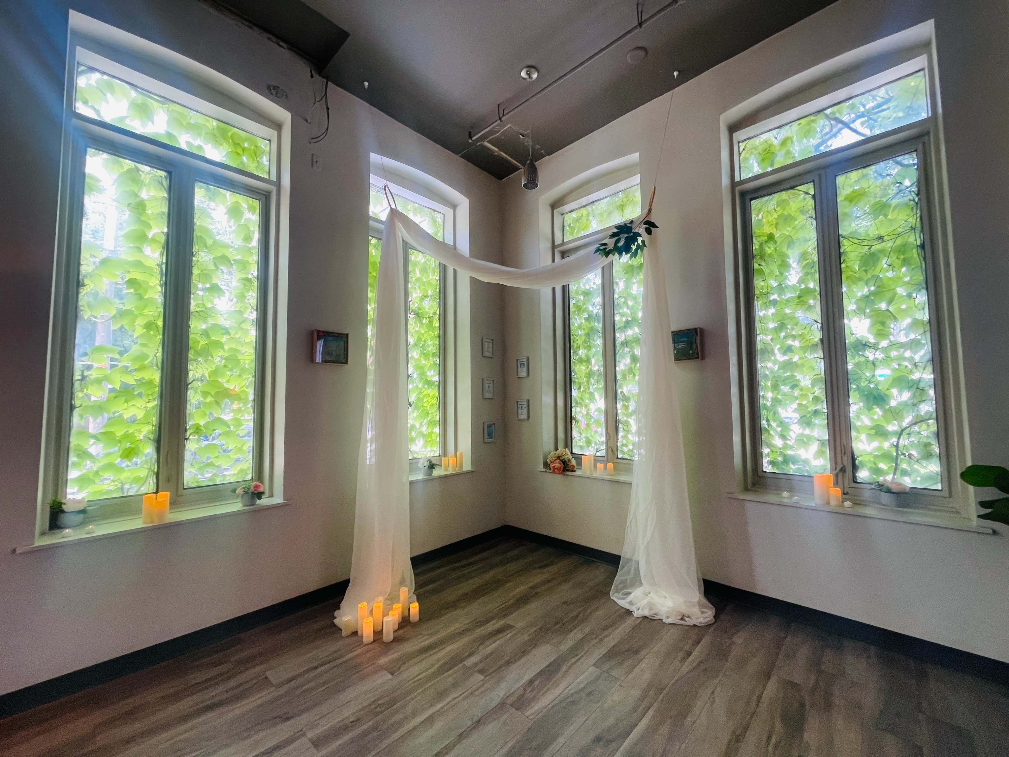 A serene room features large windows adorned with leafy greenery, softly lit by candles arranged at the base.