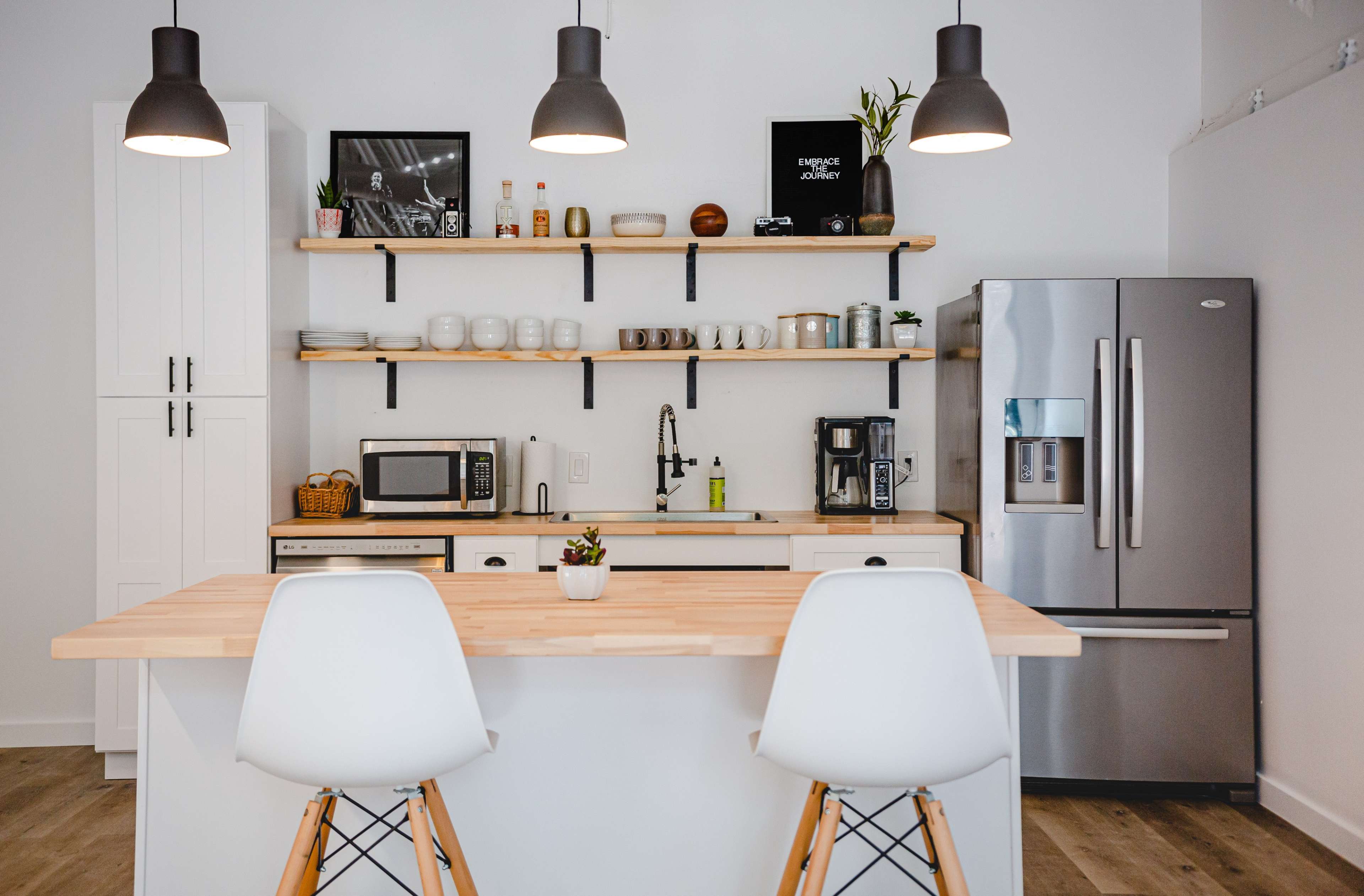 The kitchen features sleek white cabinets, a wooden countertop with two stools, open shelving displaying dishware, and modern appliances including a refrigerator and microwave.