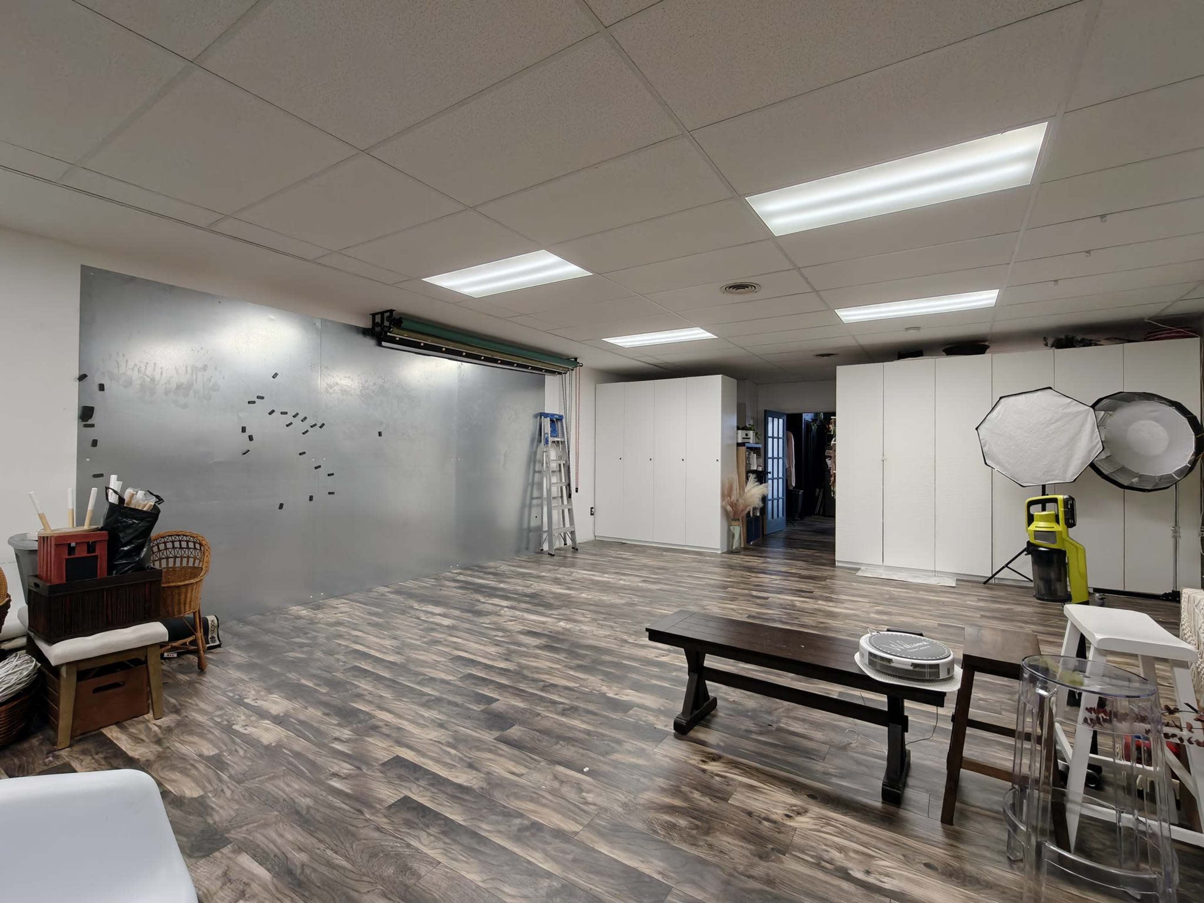 The image shows a spacious, empty studio with a polished wooden floor, a large gray wall, a folding ladder, and various storage cabinets.