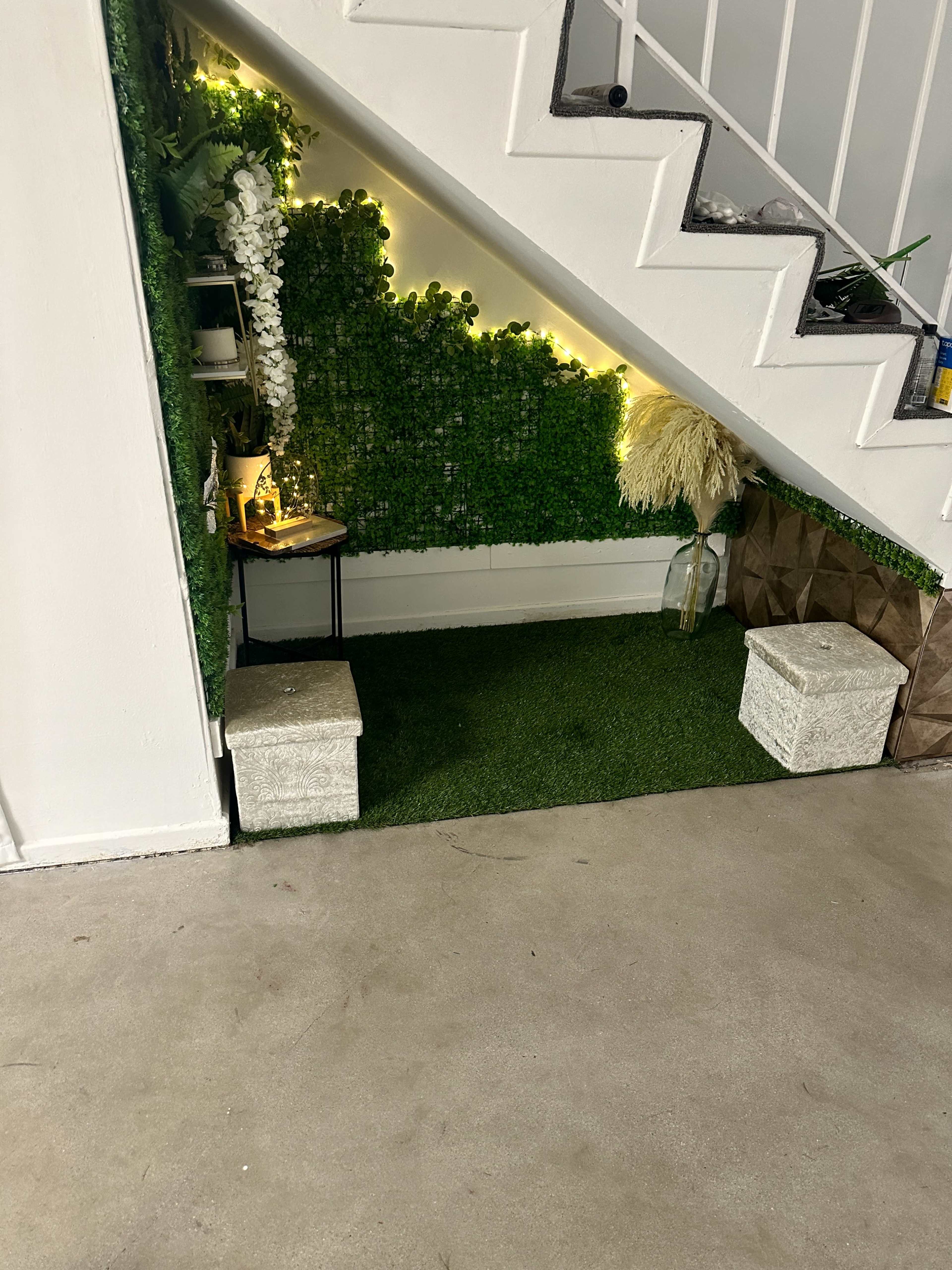 A decorative space under a staircase featuring artificial greenery, a small table with candles, and two stone-like stools.