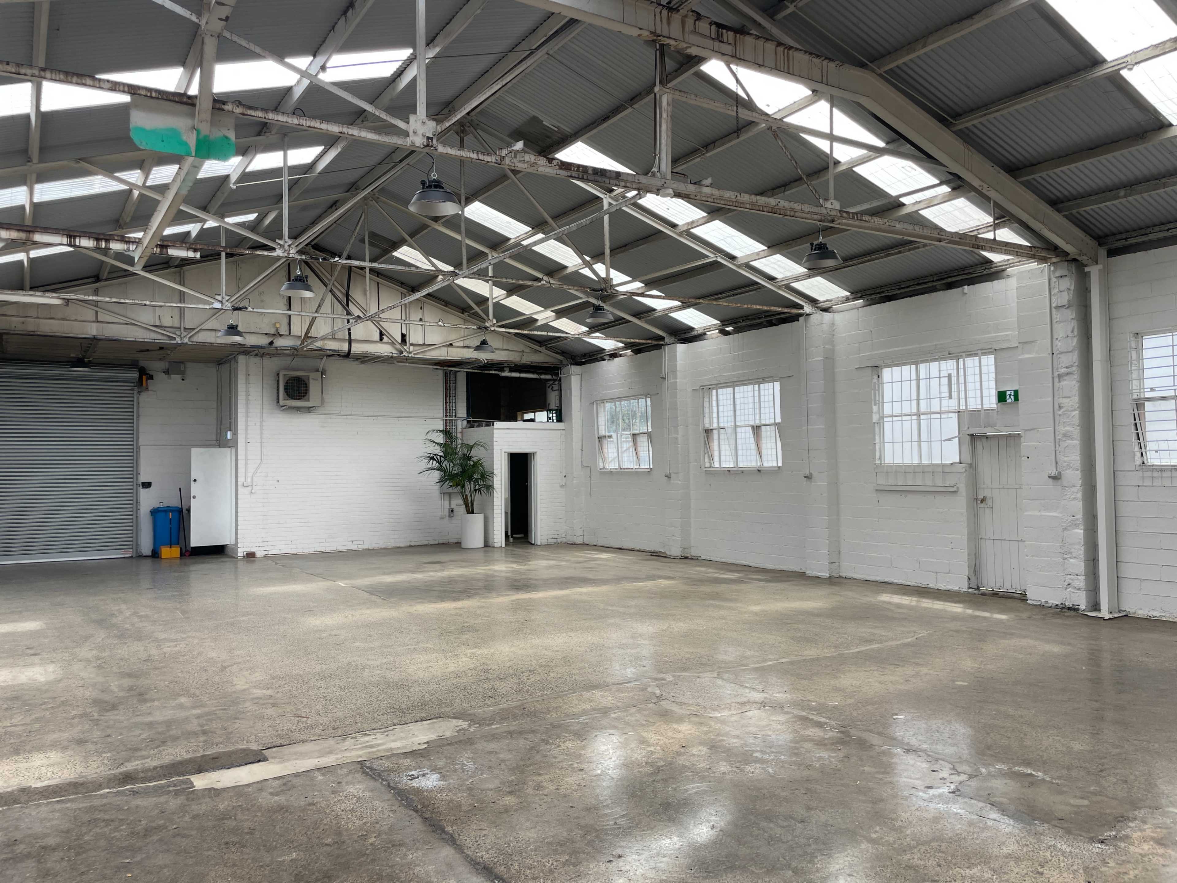 The image shows an empty industrial space with concrete flooring, white walls, large windows, and exposed beams.