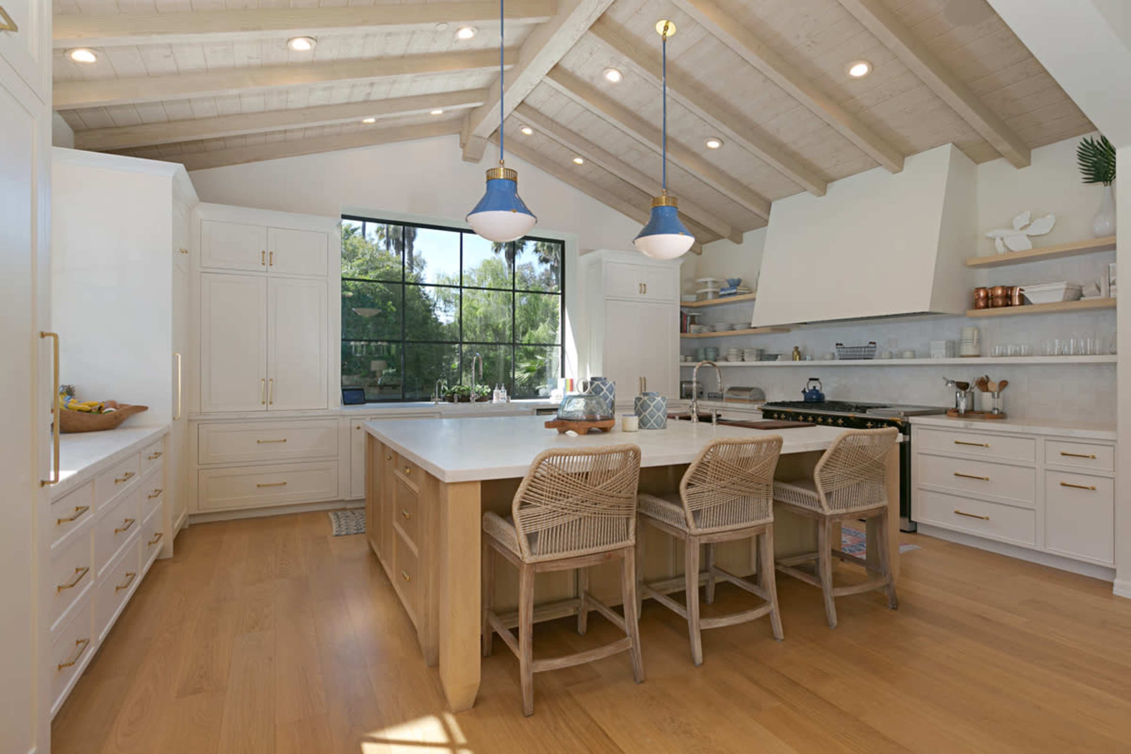 A spacious kitchen features a large island with three stools, white cabinetry, and wooden beams overhead, illuminated by pendant lights.