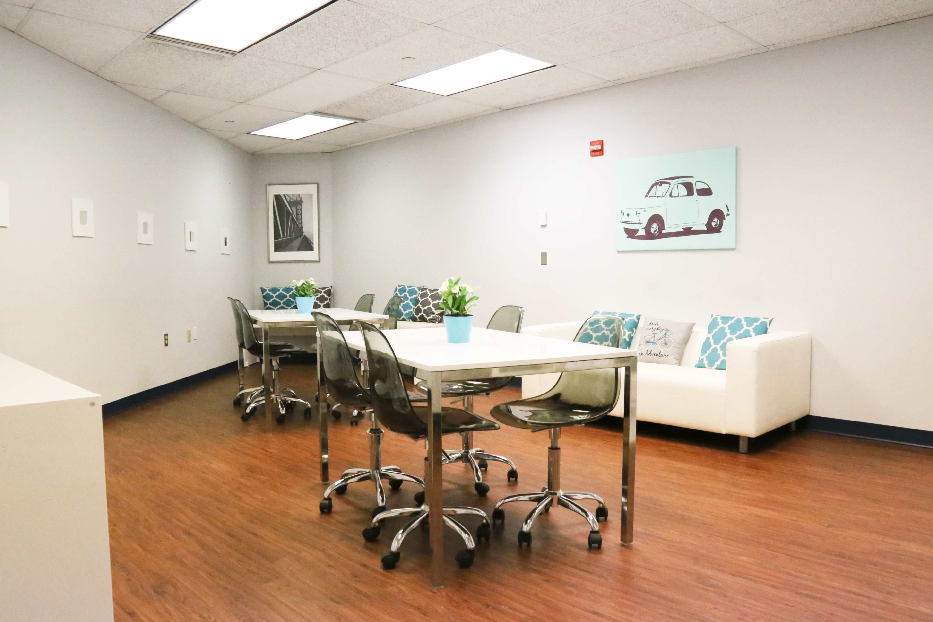 A modern office space featuring two glass tables surrounded by chairs, a white couch with decorative pillows, and wall art.