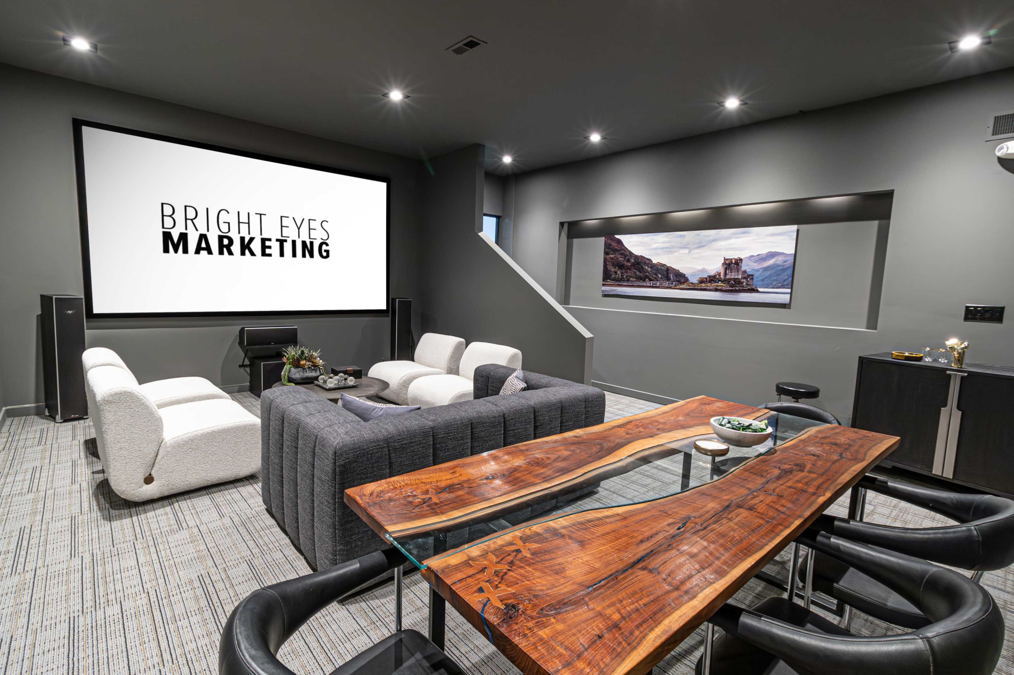 A modern conference room features a large screen displaying "BRIGHT EYES MARKETING," with a sectional sofa, a wooden table, and a contemporary design.
