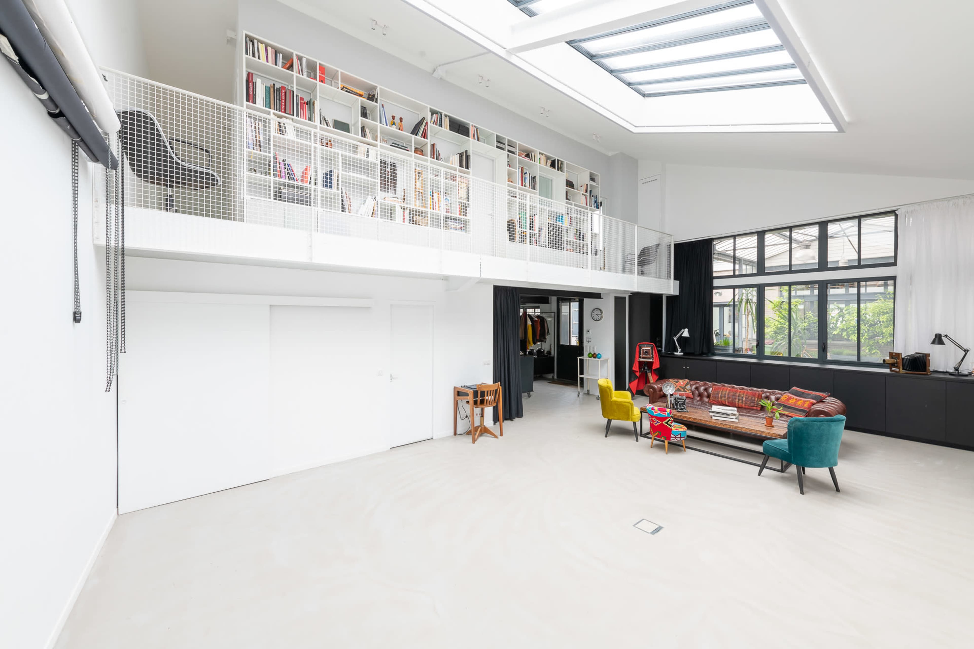 A modern, open-concept living space with a high ceiling, featuring a mezzanine library, large windows, and a colorful furniture arrangement.