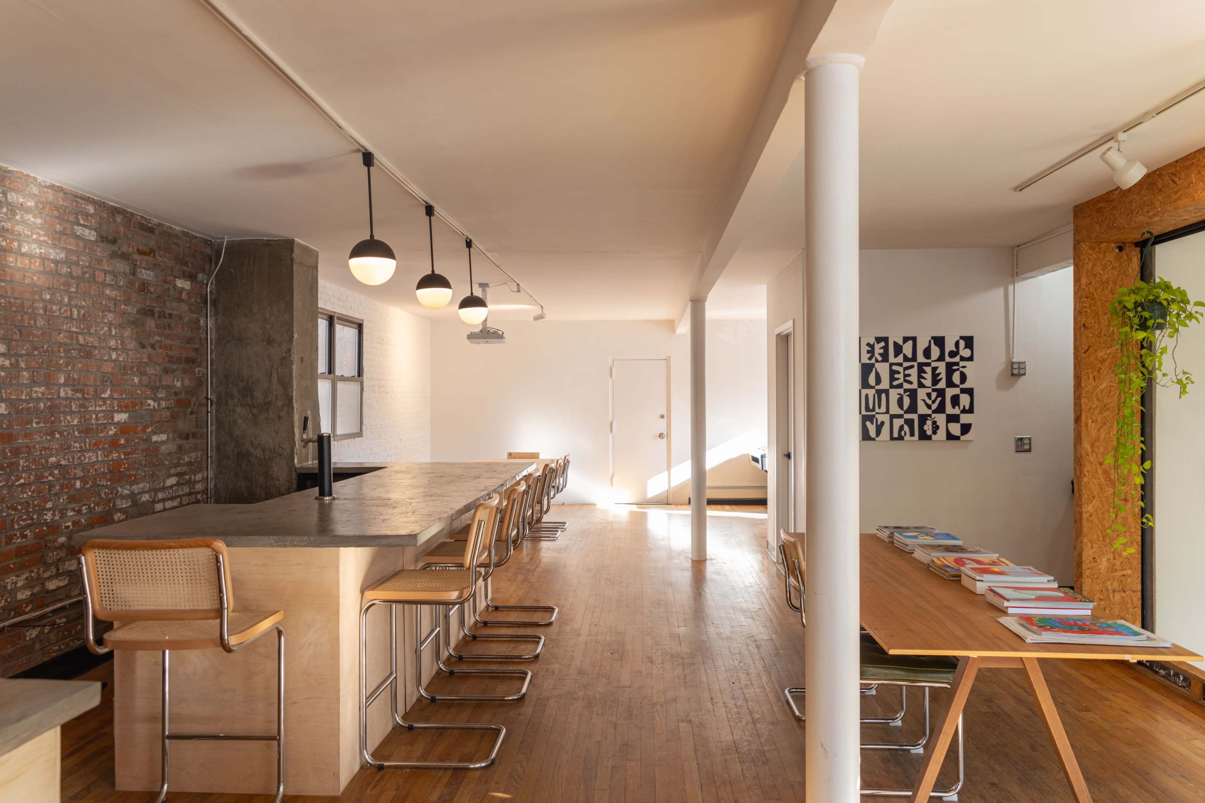 A spacious interior with a long wooden table and bar stools on one side, exposed brick walls, and a light-filled area featuring a modern design.