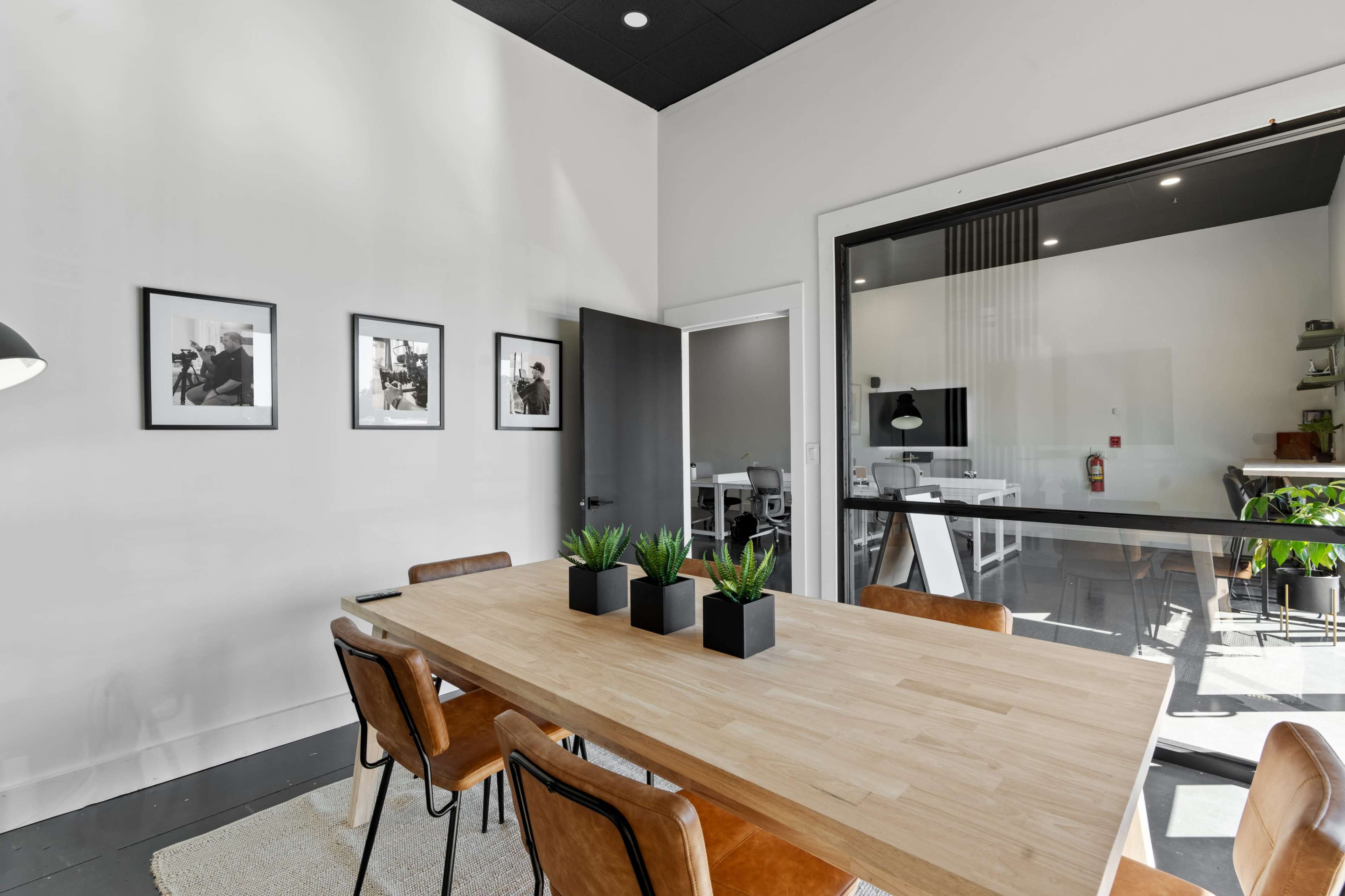 A modern conference room features a wooden table surrounded by brown chairs, with framed black-and-white photographs on the wall and a glass wall leading to another workspace.