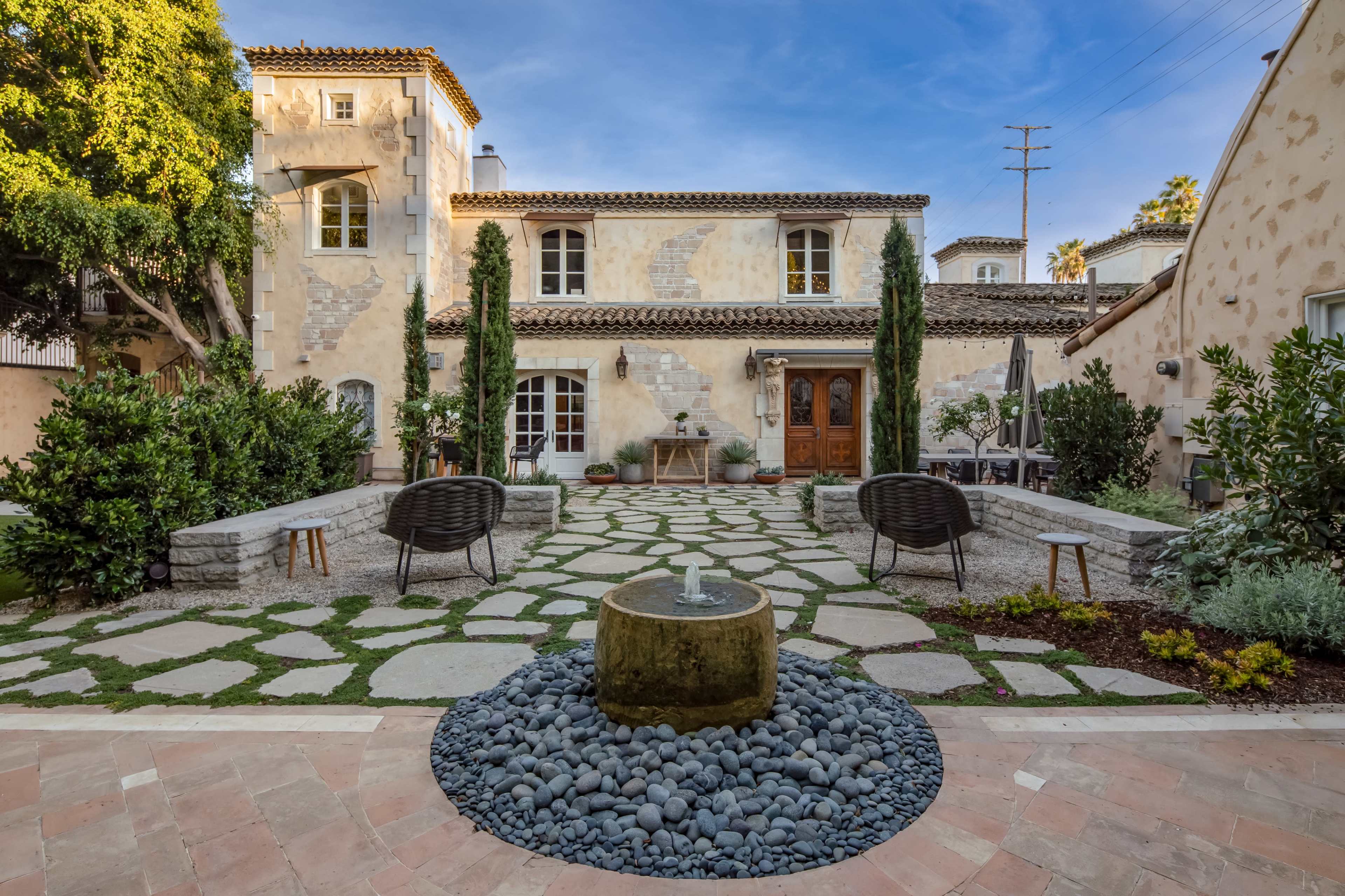 The image depicts a landscaped courtyard with stone pathways, a central water feature, and seating areas surrounded by greenery and rustic buildings.