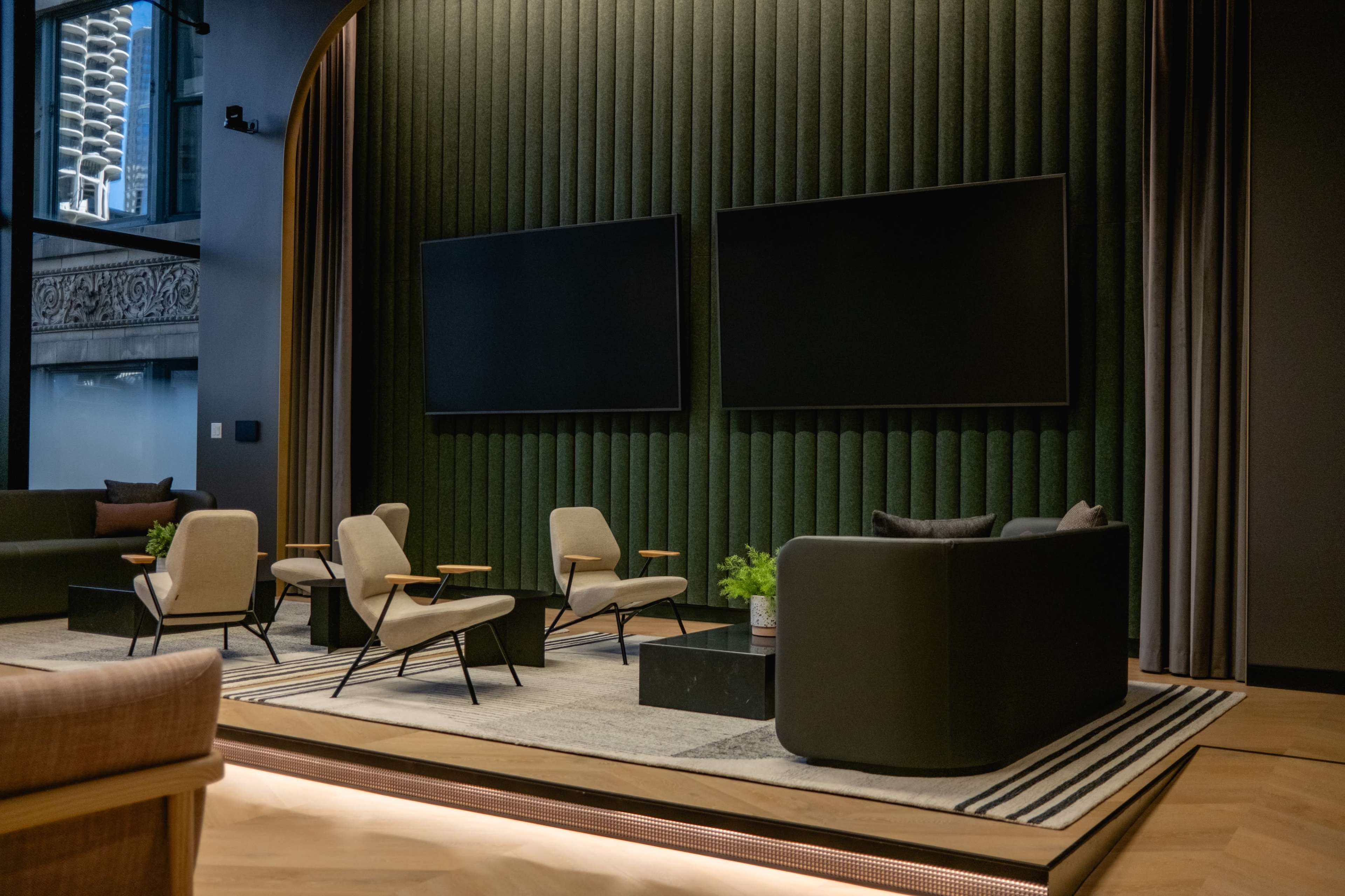 The image shows a modern lounge area with two large screens mounted on a textured green wall, surrounded by stylish seating and a patterned rug.