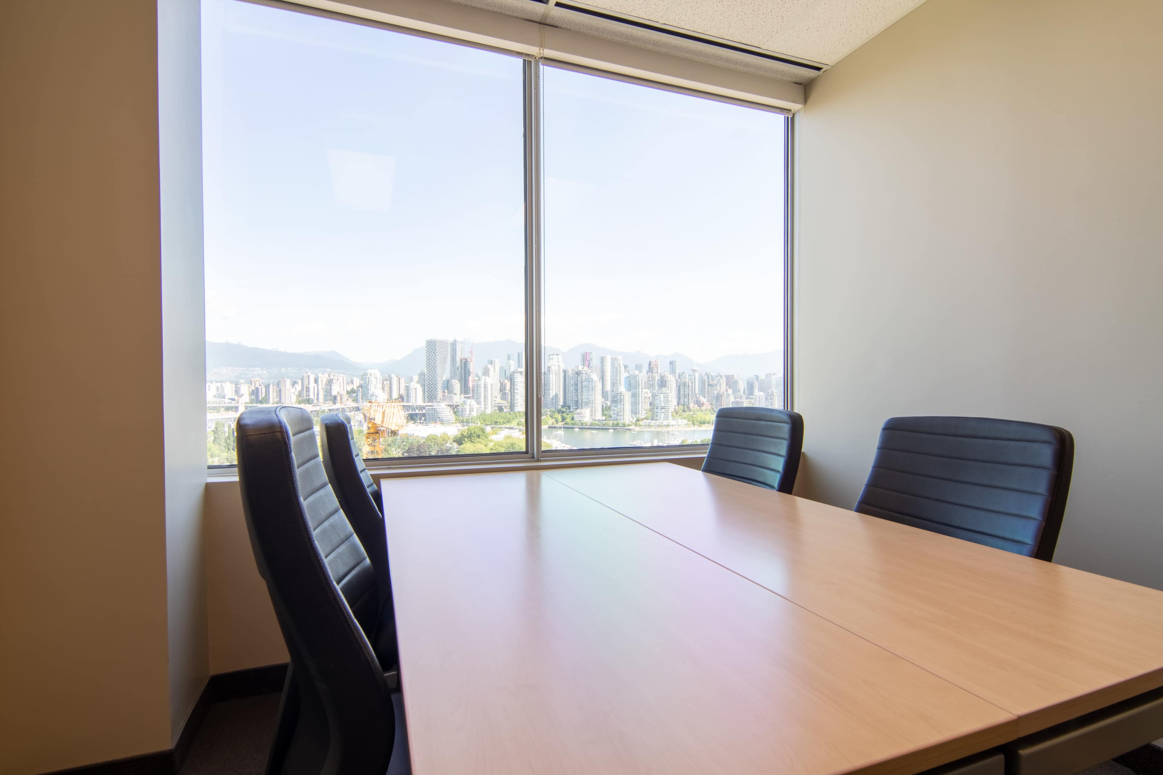 The image shows a conference room with a wooden table and black chairs, featuring a large window that offers a view of a city skyline with mountains in the background.