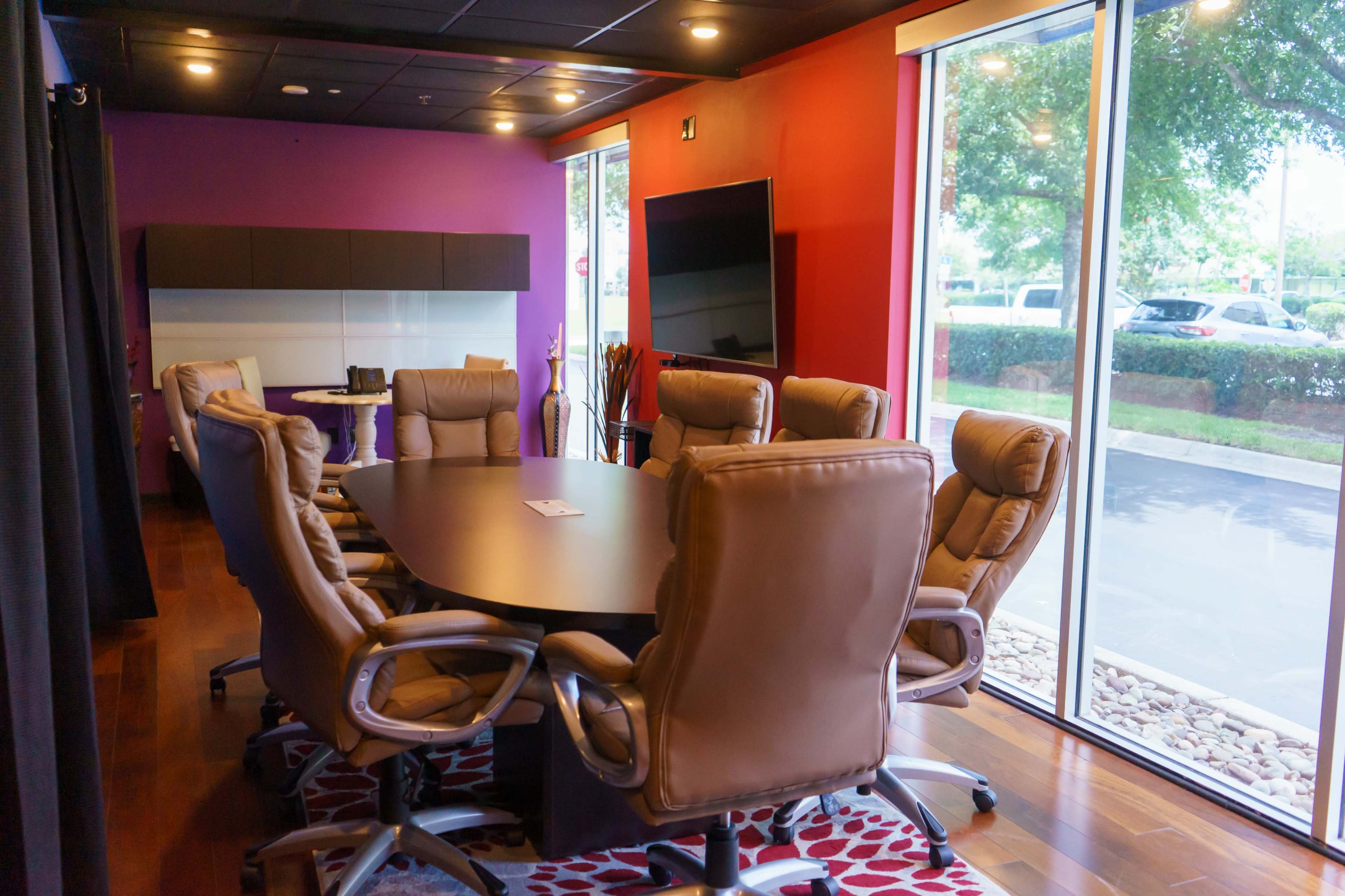 A modern conference room features a large oval table surrounded by ergonomic chairs, with a purple and red accent wall and large windows overlooking a landscaped area.