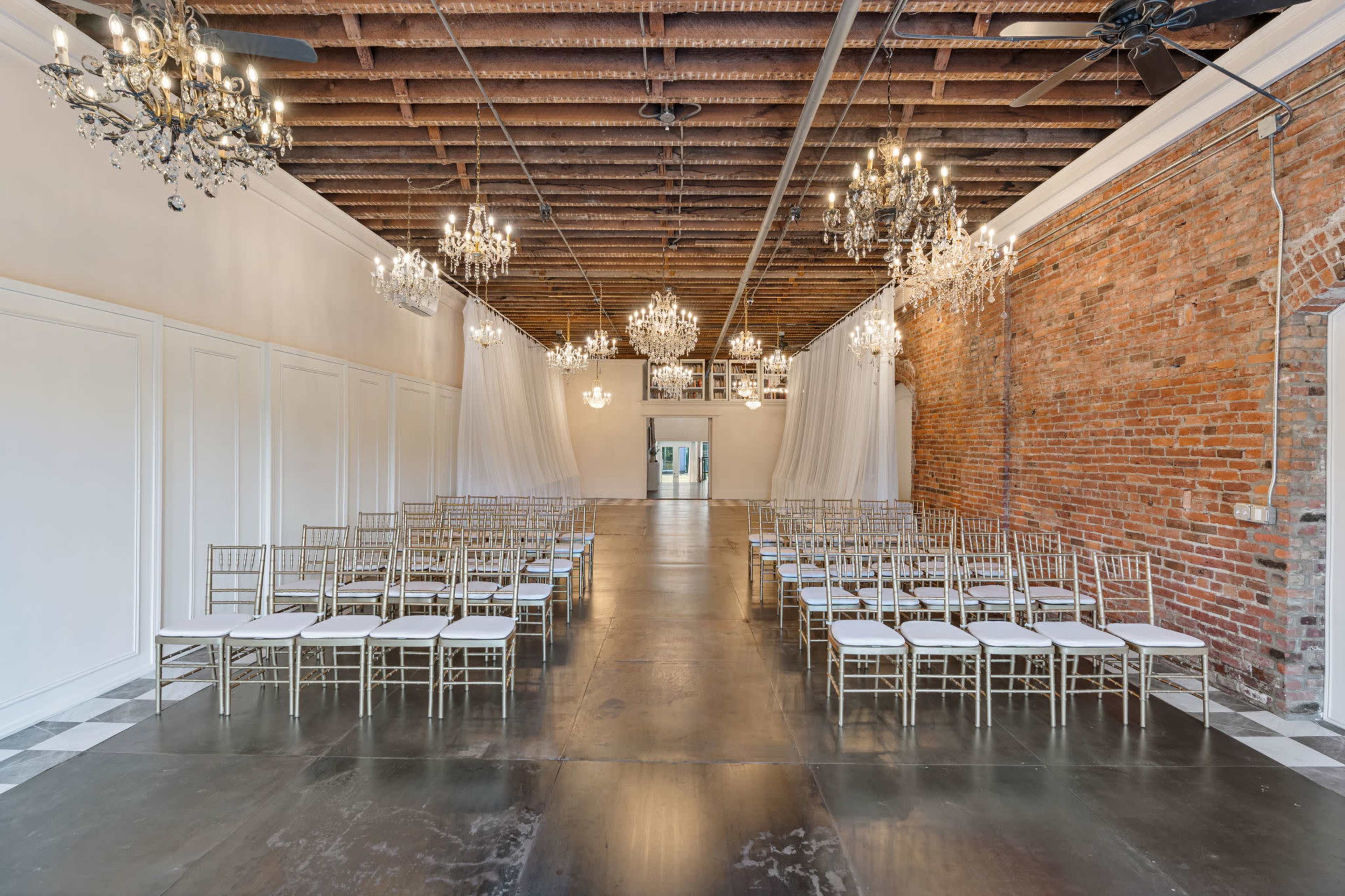 The image shows a spacious indoor venue with exposed brick walls, elegant chandeliers, and rows of gold chairs set up for an event.