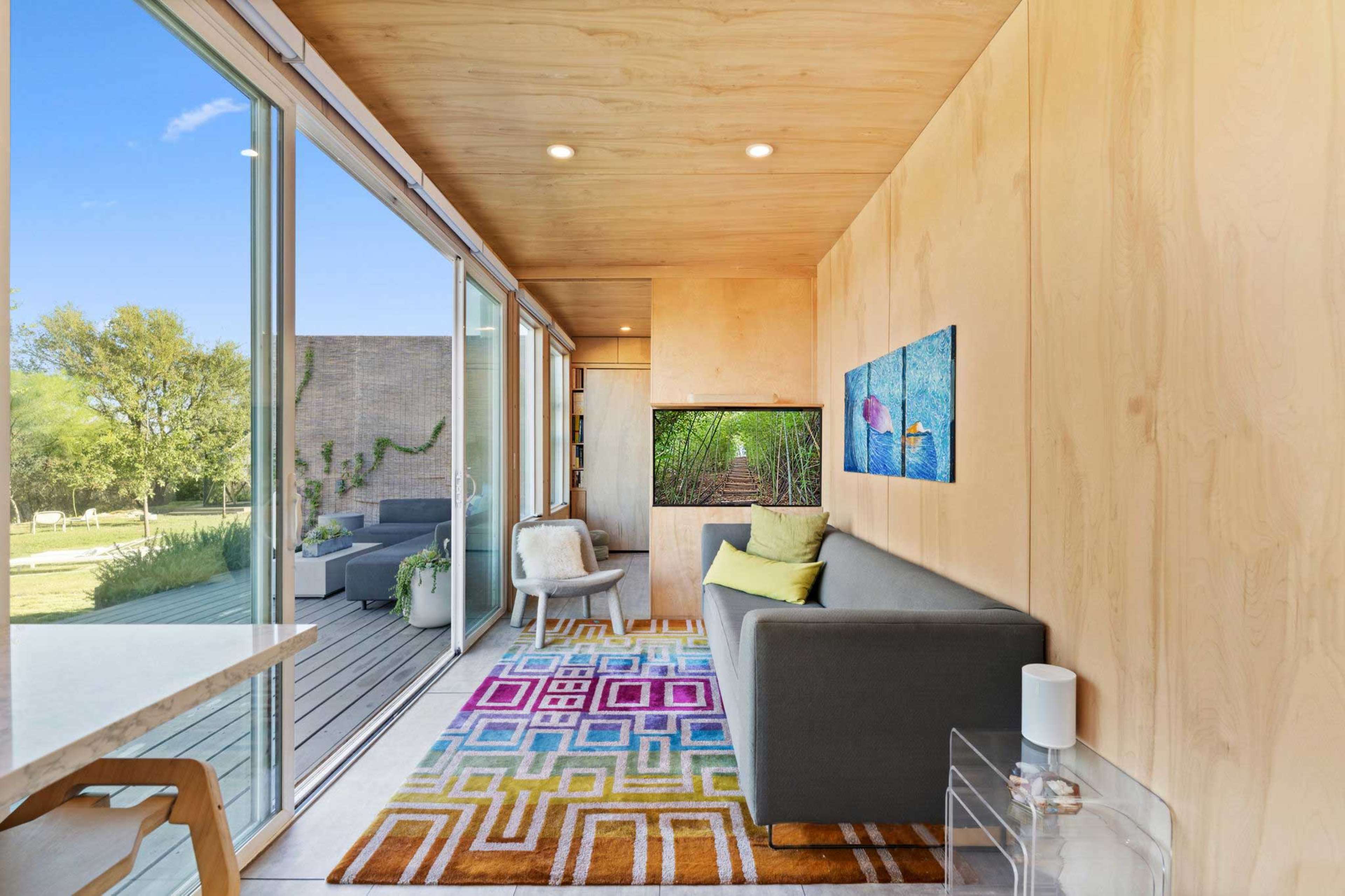 The image shows a modern interior with a plush gray sofa, a colorful area rug, and large glass windows that open to a green outdoor space.