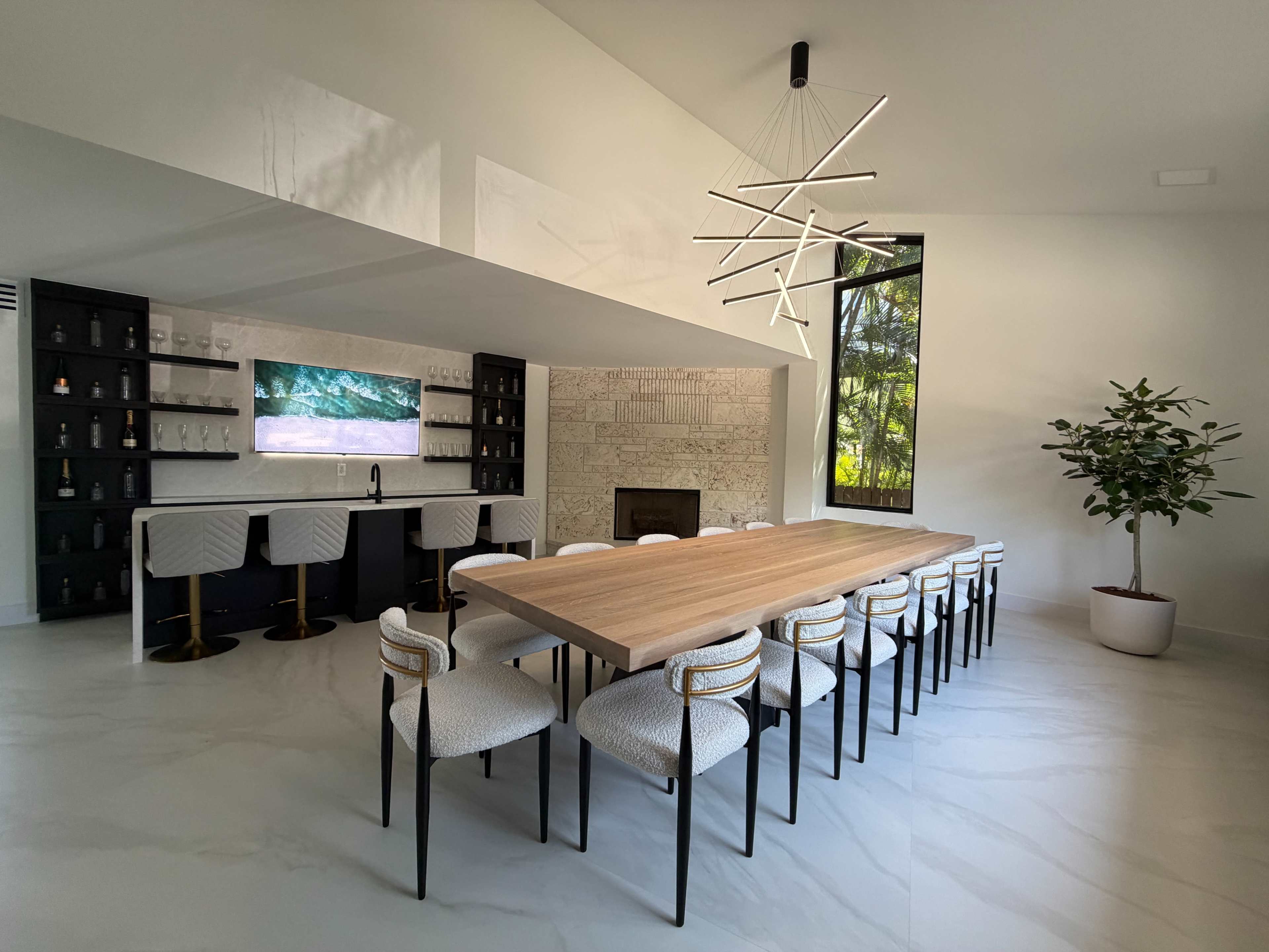 The image shows a modern dining area featuring a long wooden table with ten chairs, a sleek bar with a screen, a fireplace, and large windows that let in natural light.
