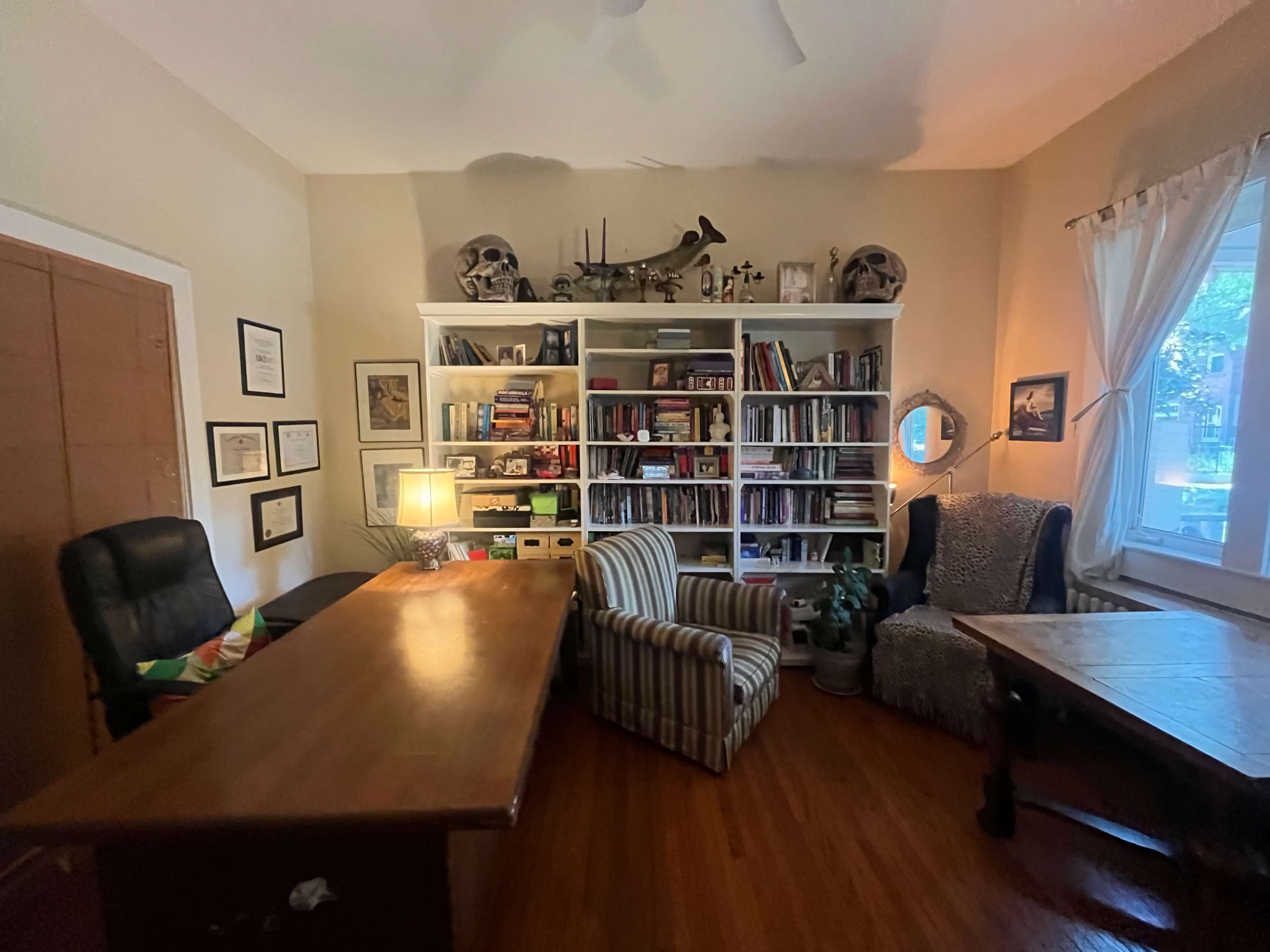 The image shows a cozy office space featuring a large wooden desk, a striped armchair, a bookshelf filled with books and decorative items, and a window with curtains.