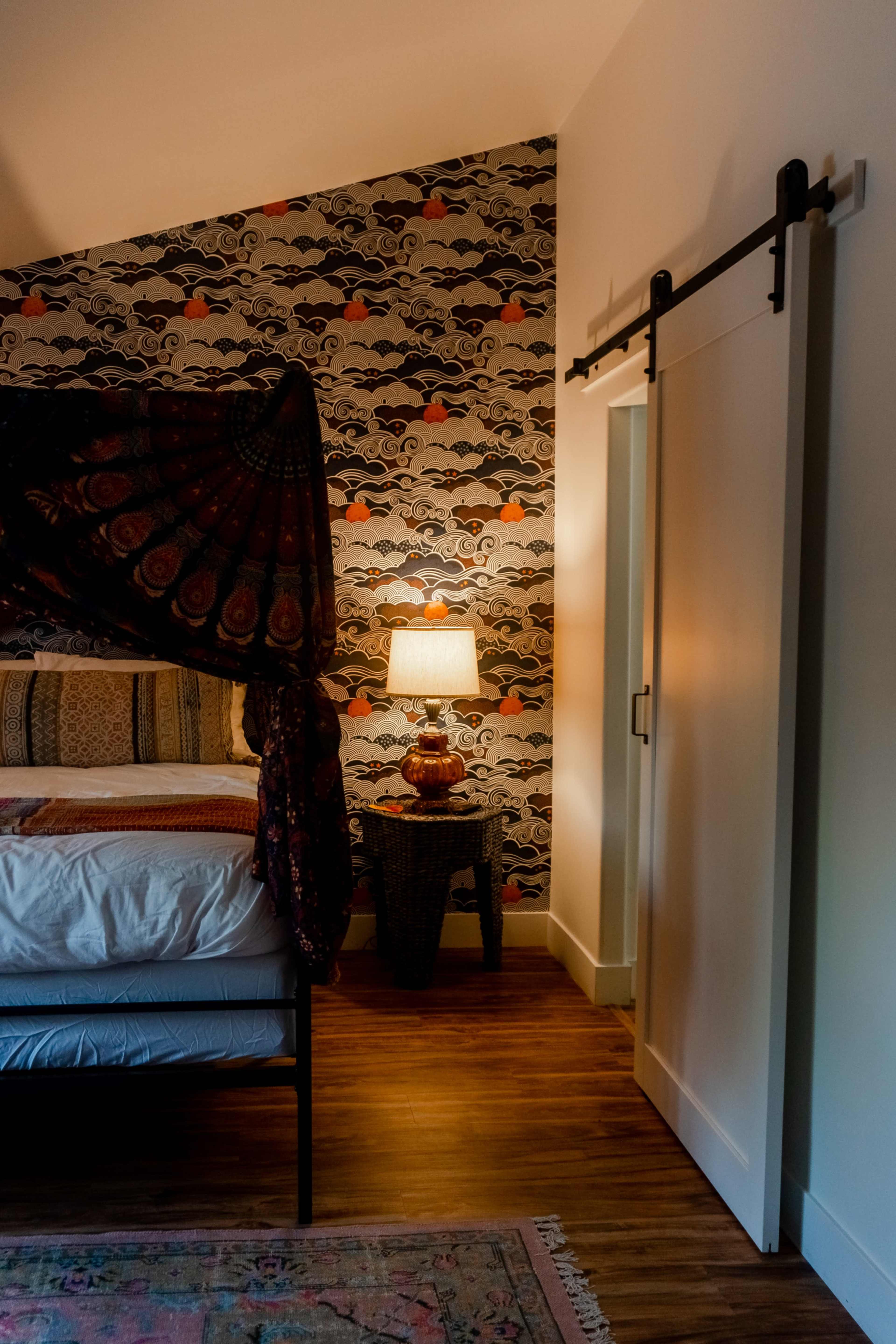 A bedroom with a patterned wallpaper in the background, a bed with a canopy, a lamp on a side table, and a sliding door.
