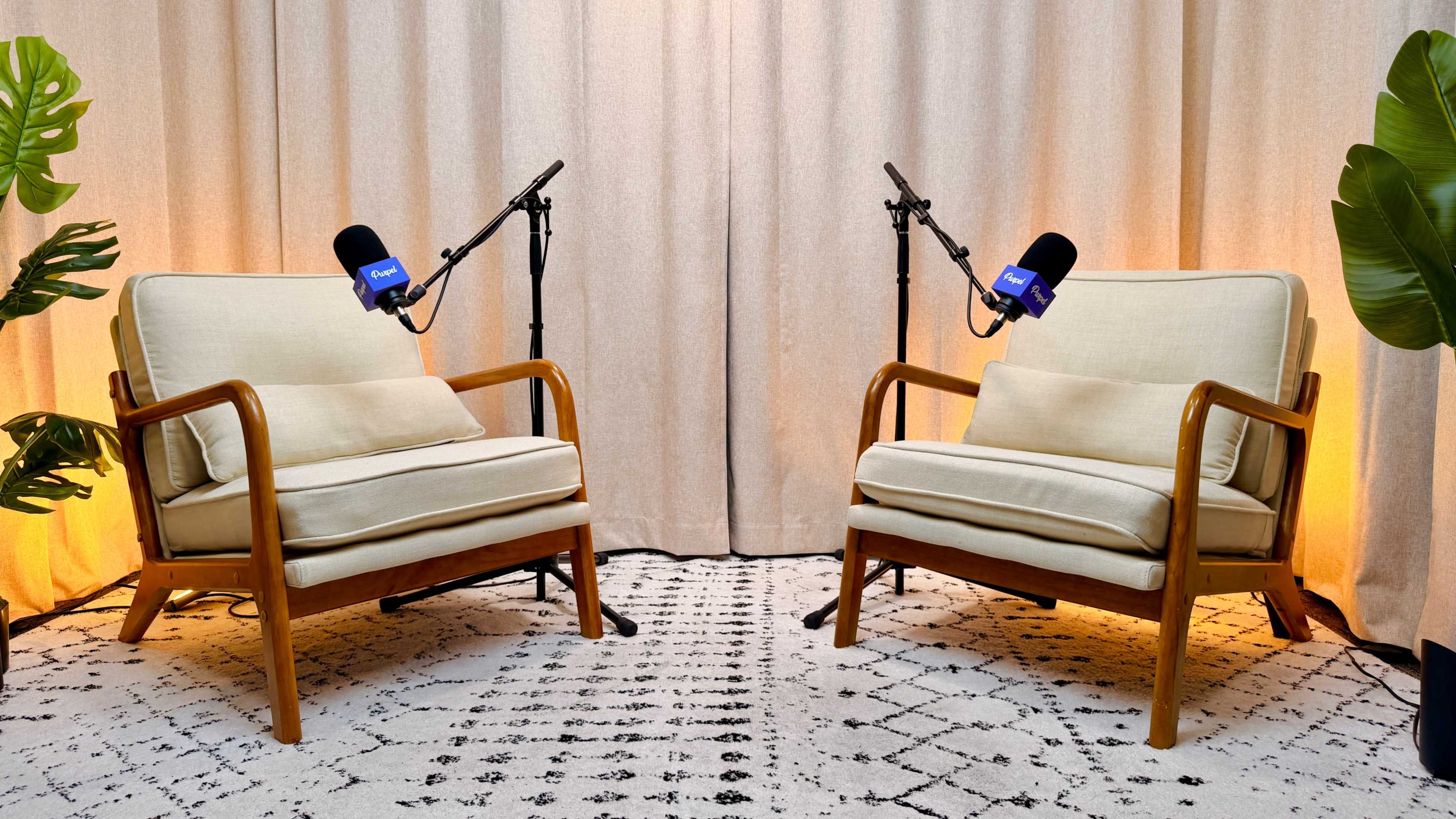 The image features two beige chairs with wooden frames positioned opposite each other, accompanied by microphone stands on either side, against a backdrop of curtains and soft lighting.