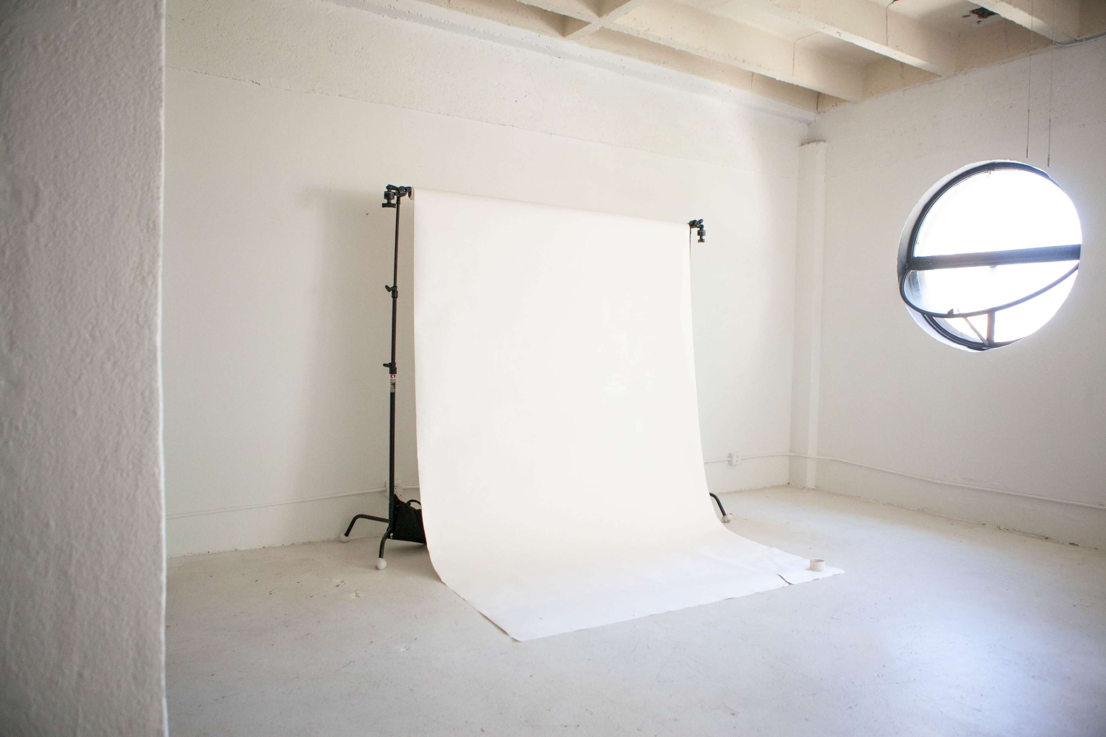 The image shows a spacious, empty room with a white backdrop set up on a stand in front of a circular window.