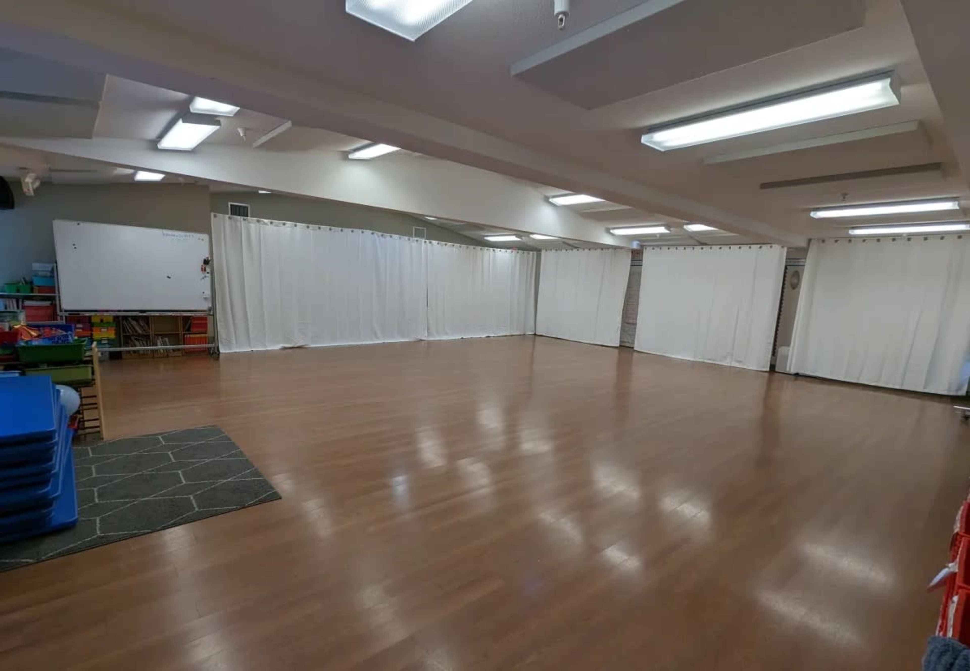 The image shows a spacious indoor room with wooden floors, white curtains dividing the space, and neatly stacked blue mats in one corner.