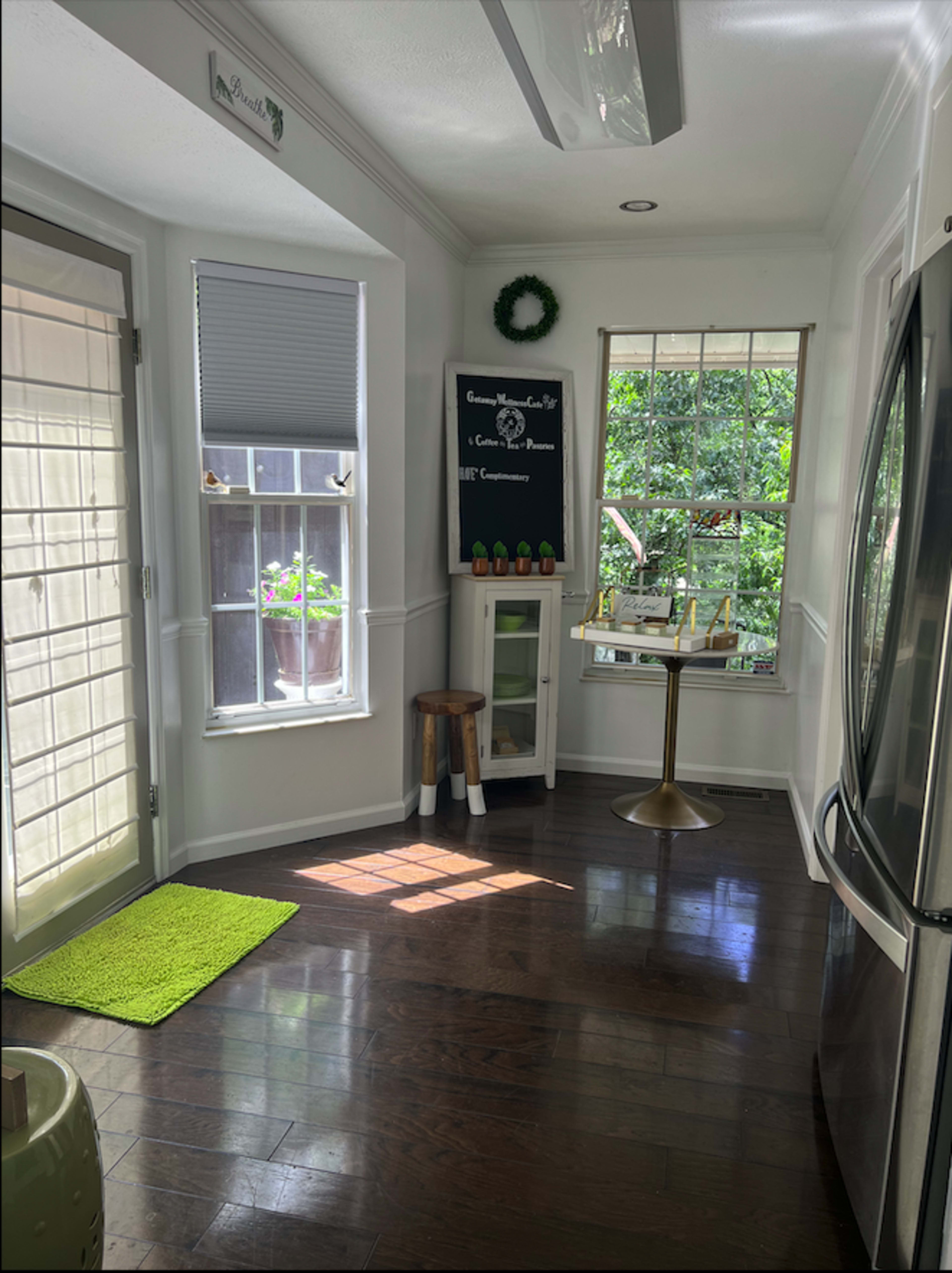 A bright kitchen nook features a glass door, large windows with plants, a small table, and a refrigerator.