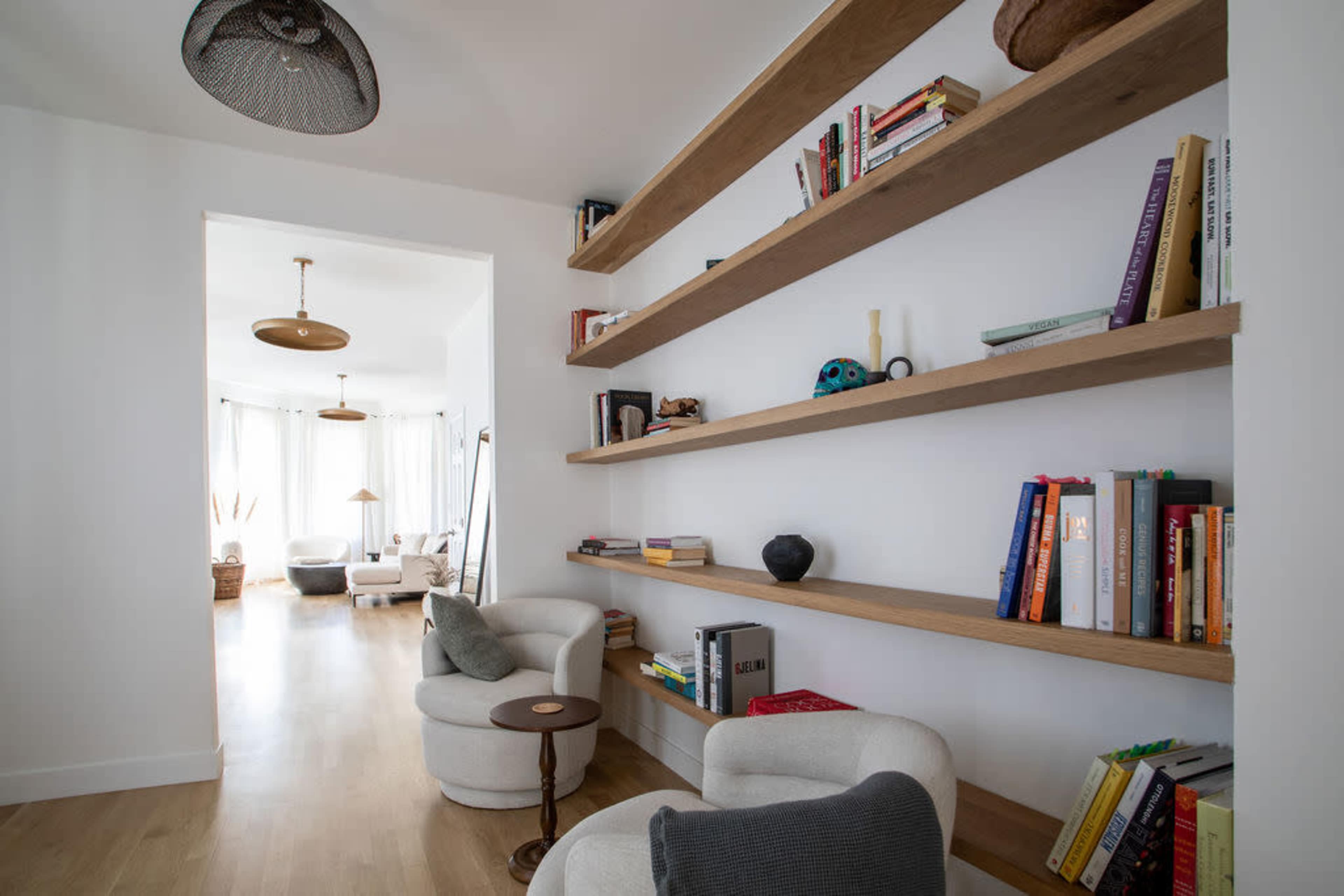 A modern interior featuring wooden shelves filled with books, two white armchairs, and a view into a well-lit living area.