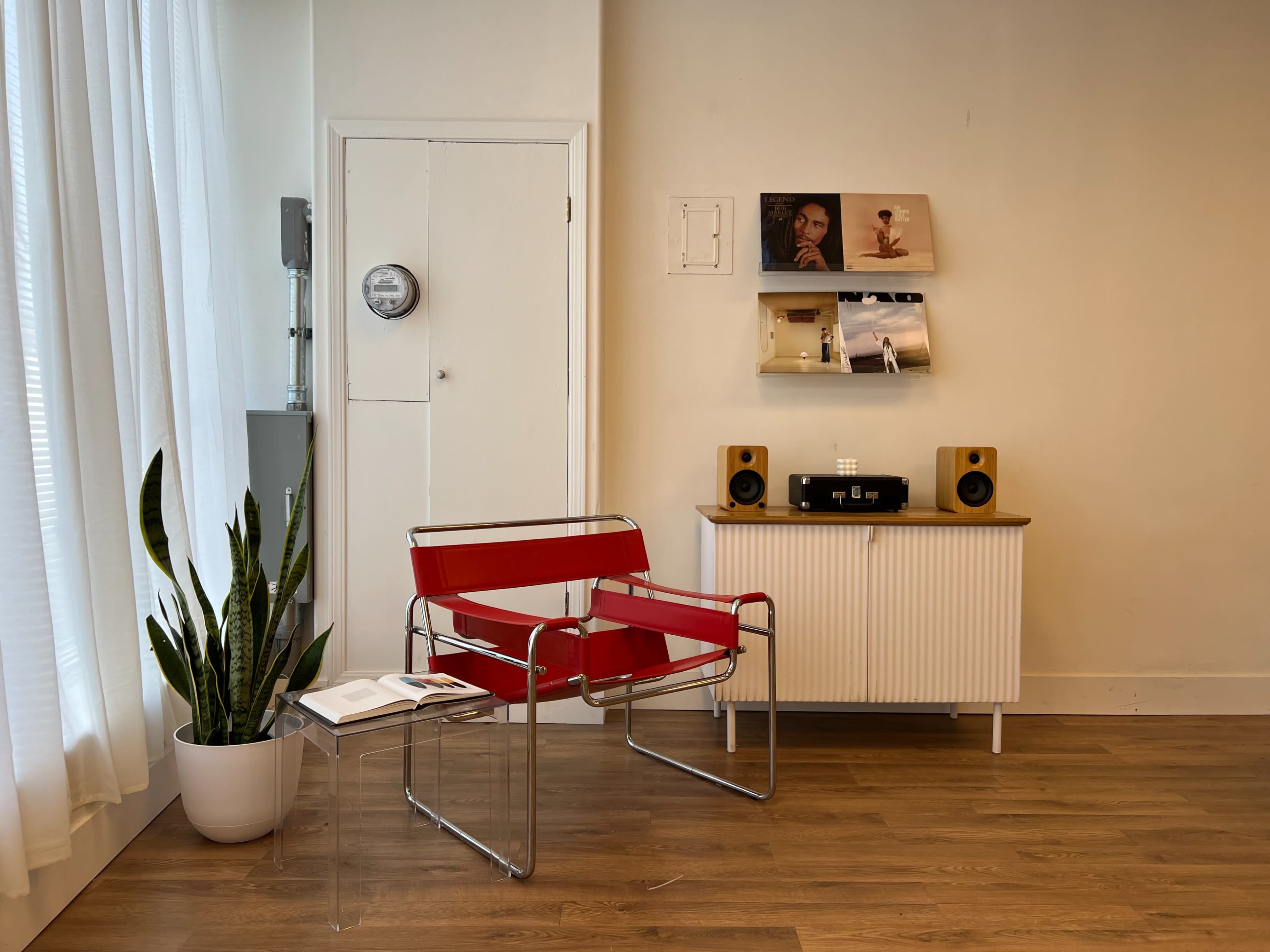 The image shows a modern interior with a red chair, a white cabinet with speakers, and a plant near large windows.