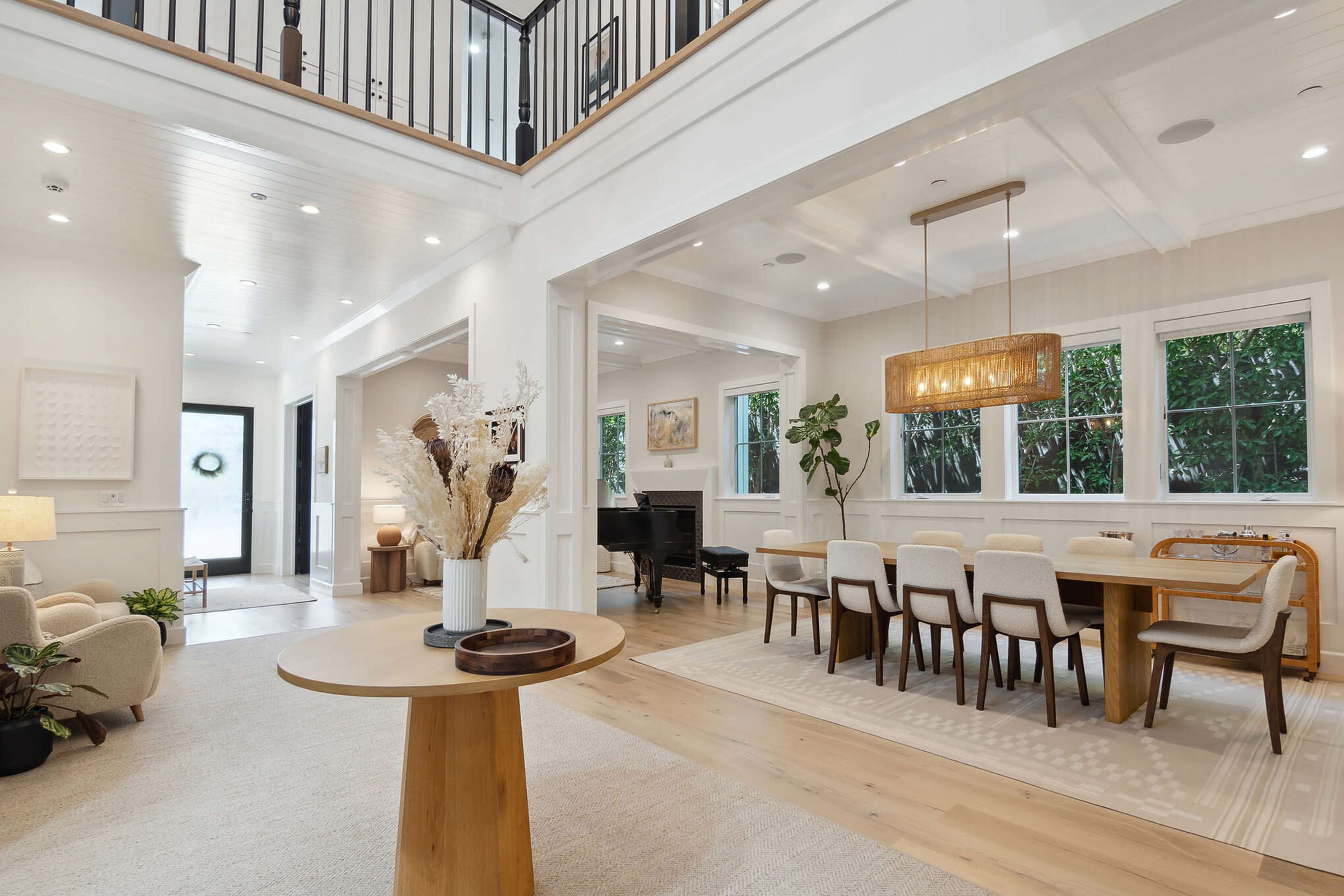 A spacious, modern living area that includes a dining area with a large table and a piano, framed by large windows and light-colored furnishings.