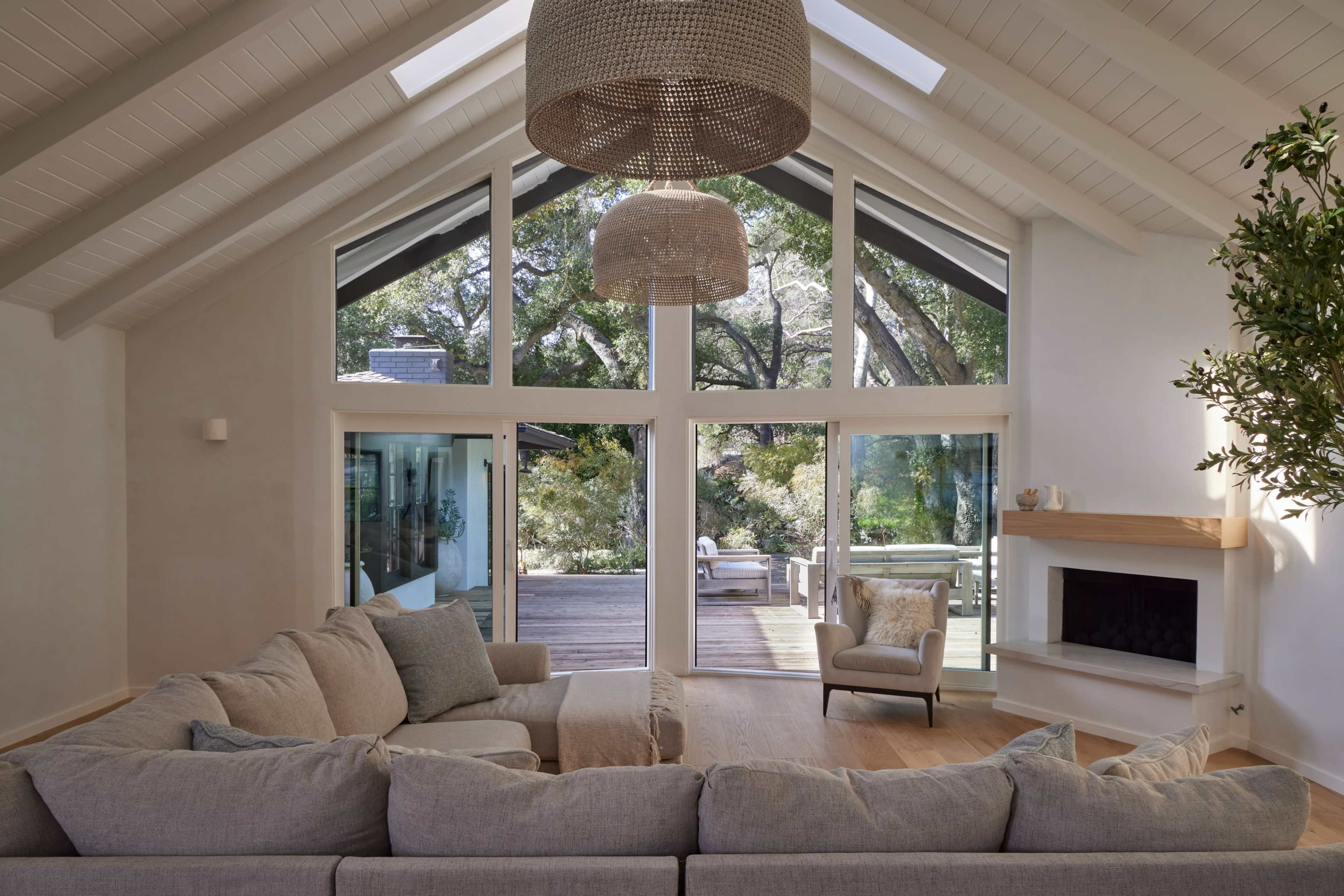 The image depicts a spacious living room with large windows overlooking a deck and garden, featuring a light-colored sofa and a modern light fixture.