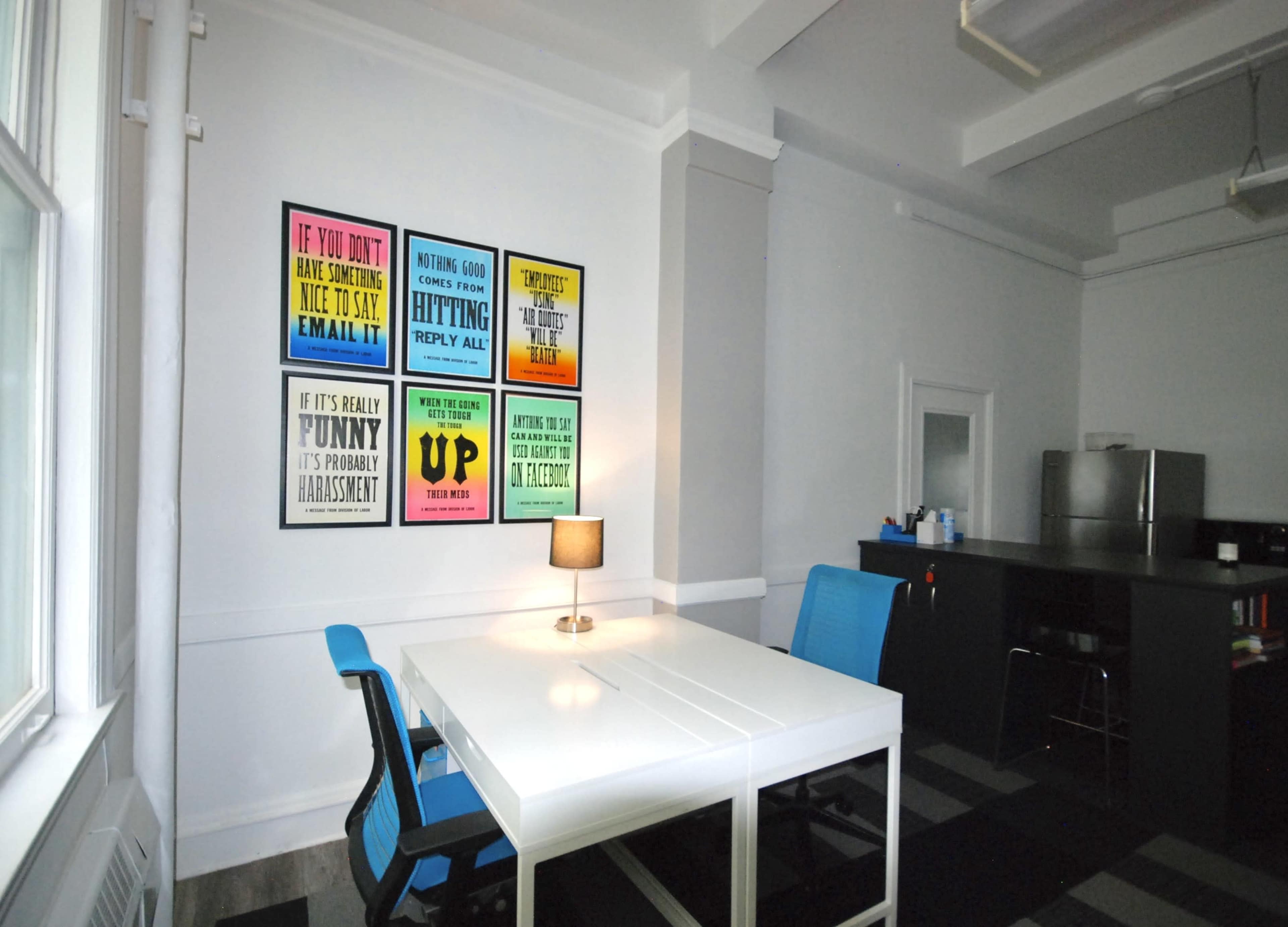 The image shows a minimalist office space with a white table, two blue chairs, and colorful motivational posters on the wall.