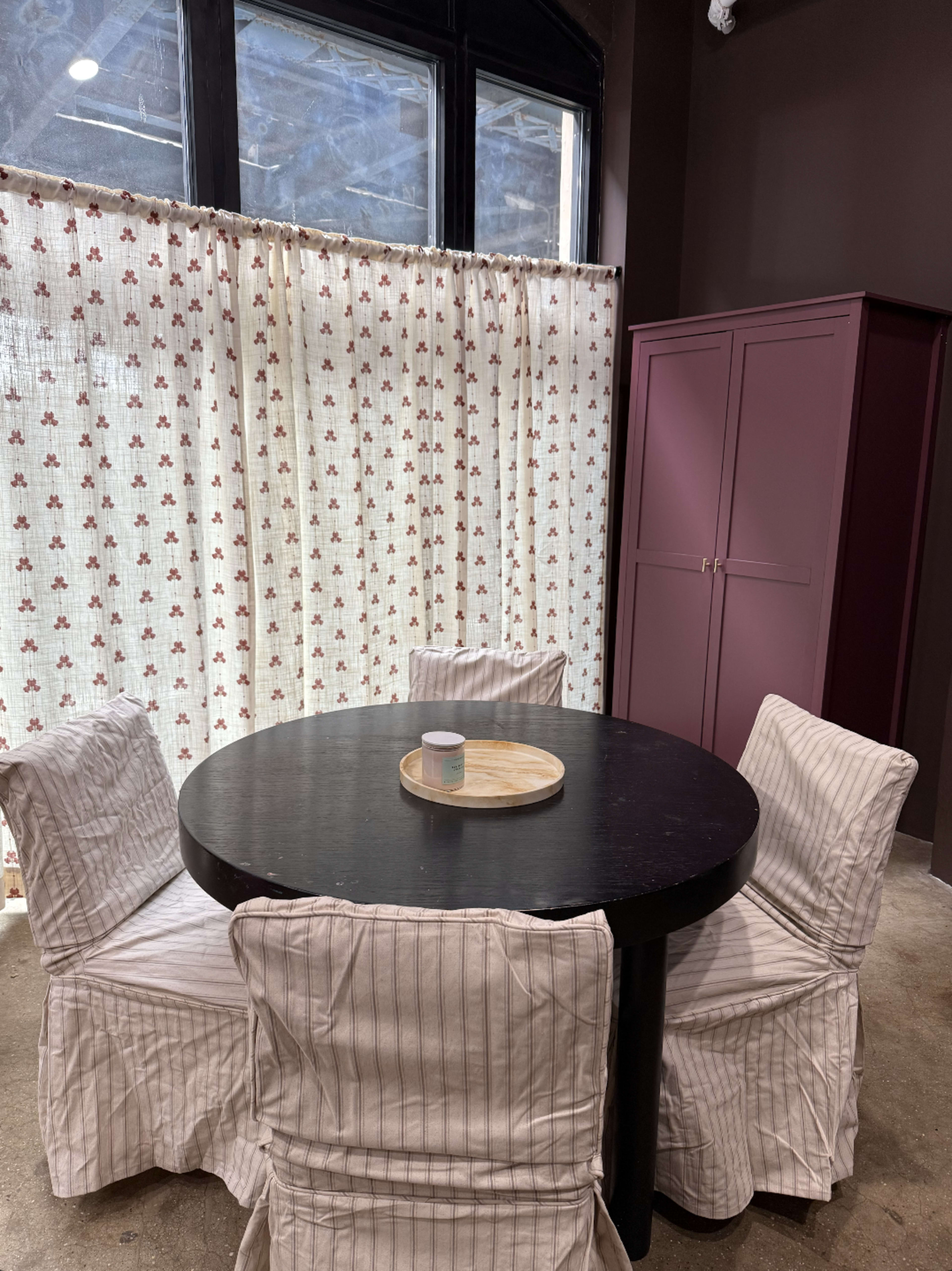 The image shows a round black table surrounded by four upholstered chairs draped in striped fabric, with a wooden tray in the center, near a curtained window and a purple cabinet.
