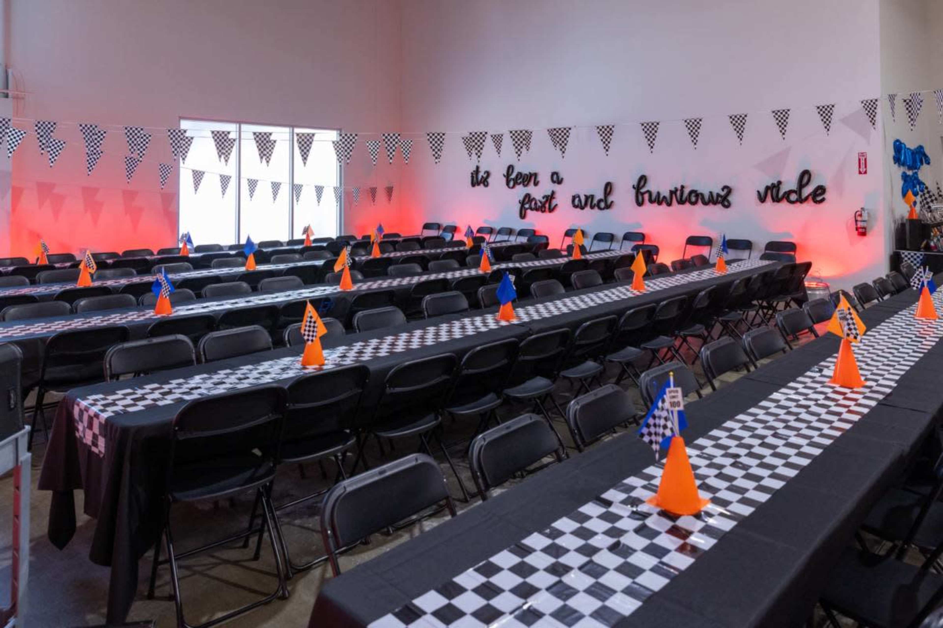 The image shows a large indoor space set up for a themed event with rows of black tables covered with checkered tablecloths, each featuring orange cones with blue and orange flags.