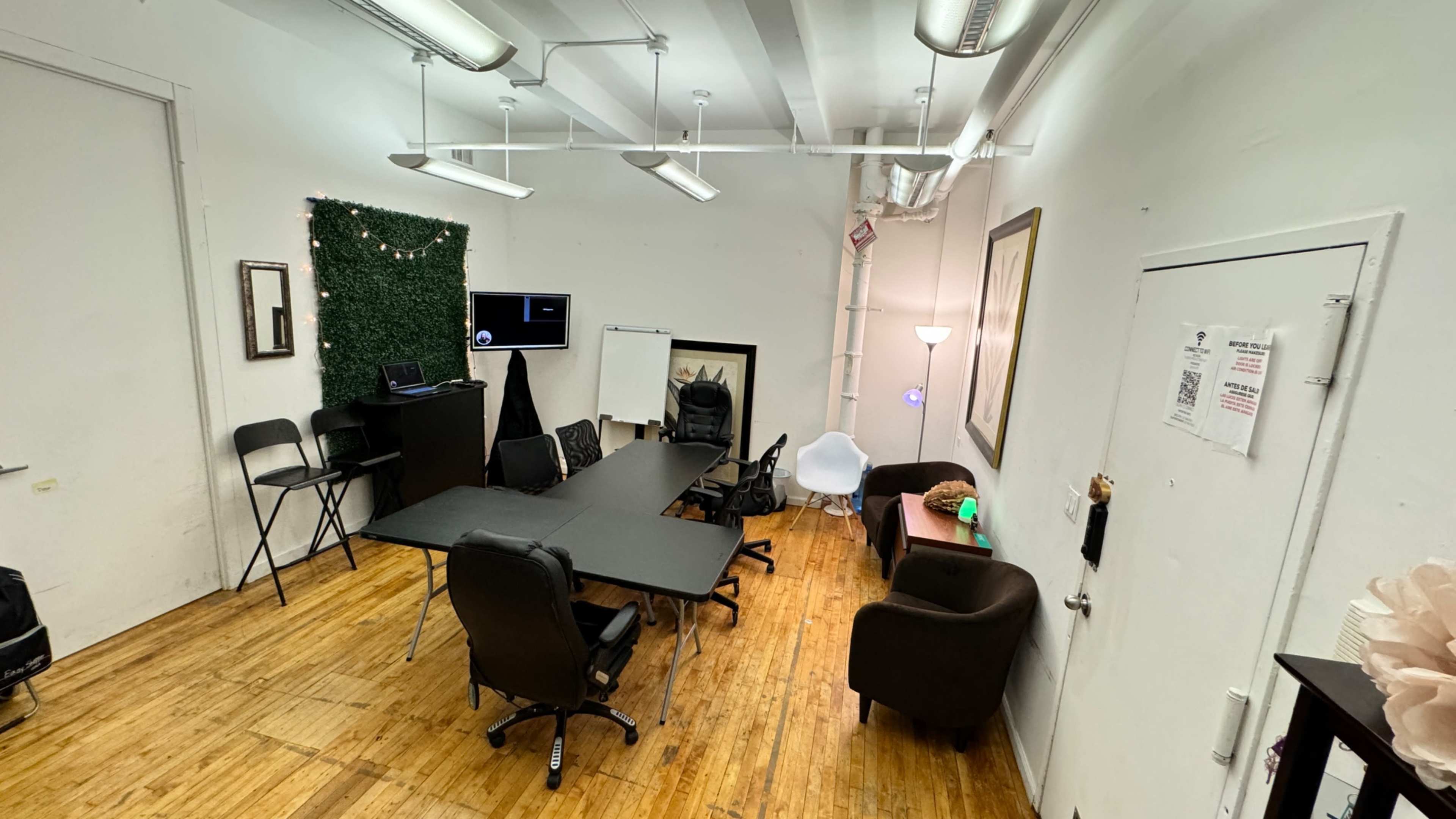 A conference room with a large black table surrounded by office chairs, a whiteboard, a television screen mounted on the wall, and some seating areas with a lamp.