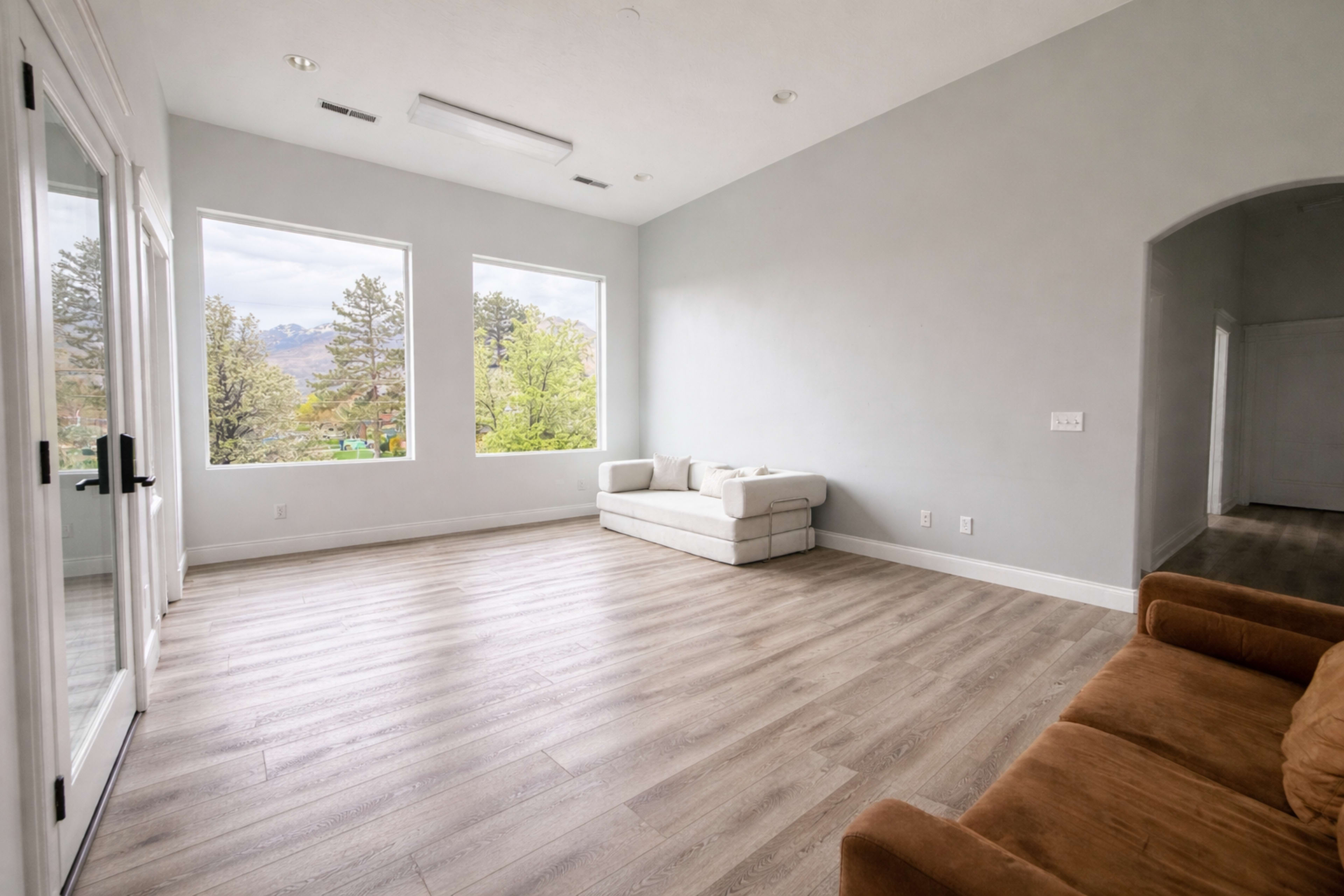 The image shows a spacious, empty room with large windows, a light-colored couch, and wooden flooring.