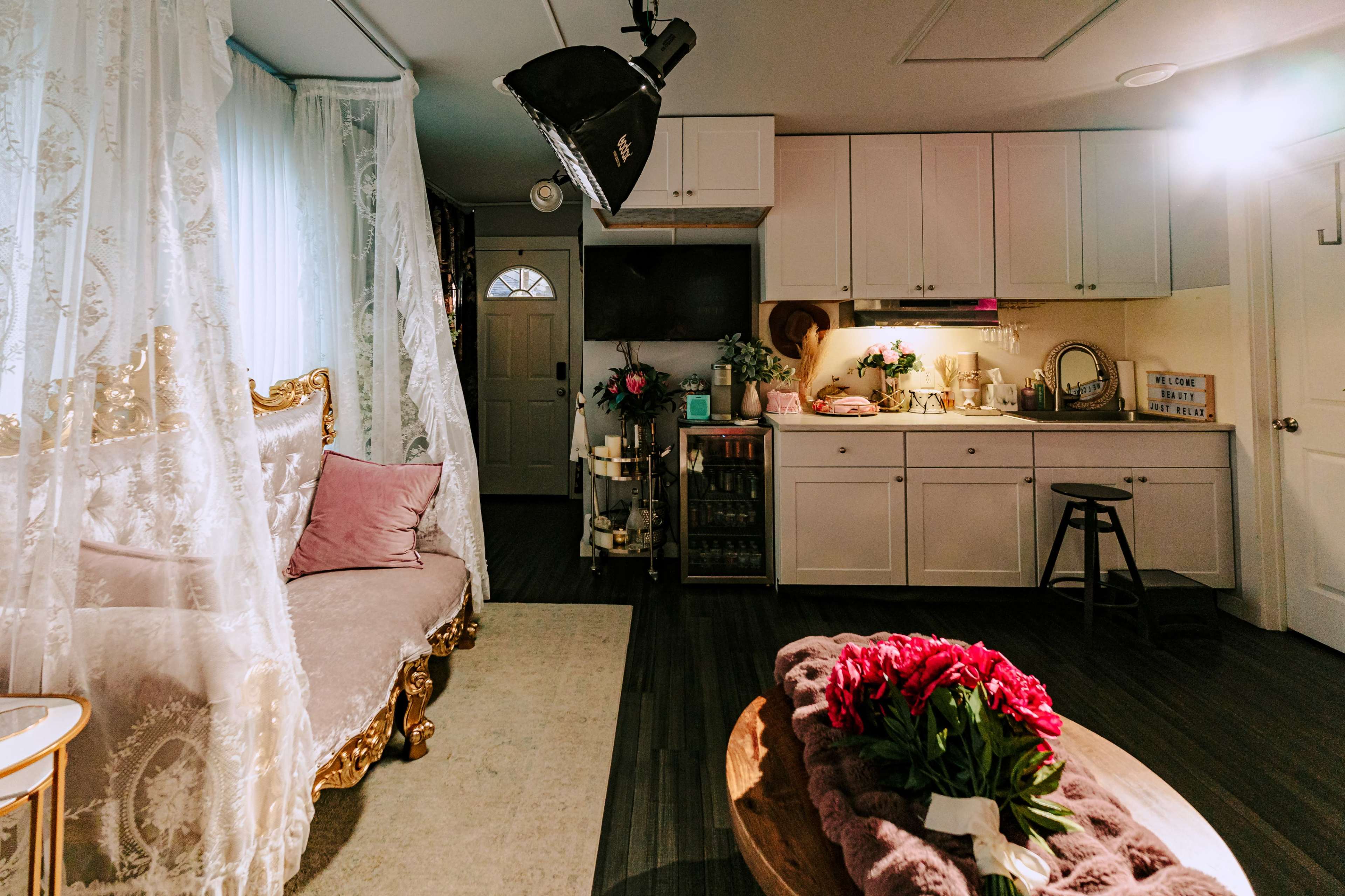 The image shows a well-decorated small room with a couch, a kitchen area, and a bouquet of flowers on a coffee table.