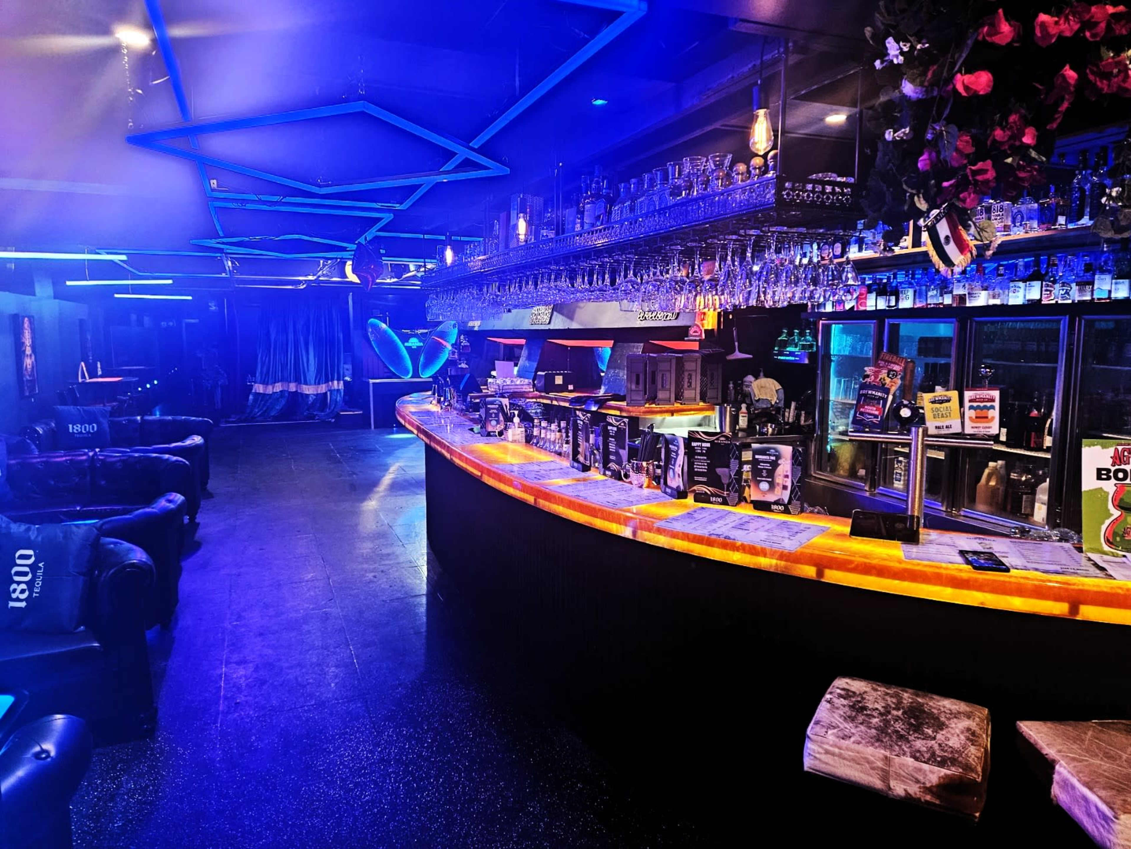 The image shows a dimly lit bar featuring a curved illuminated counter, rows of hanging glassware, and various seating options arranged around the space.