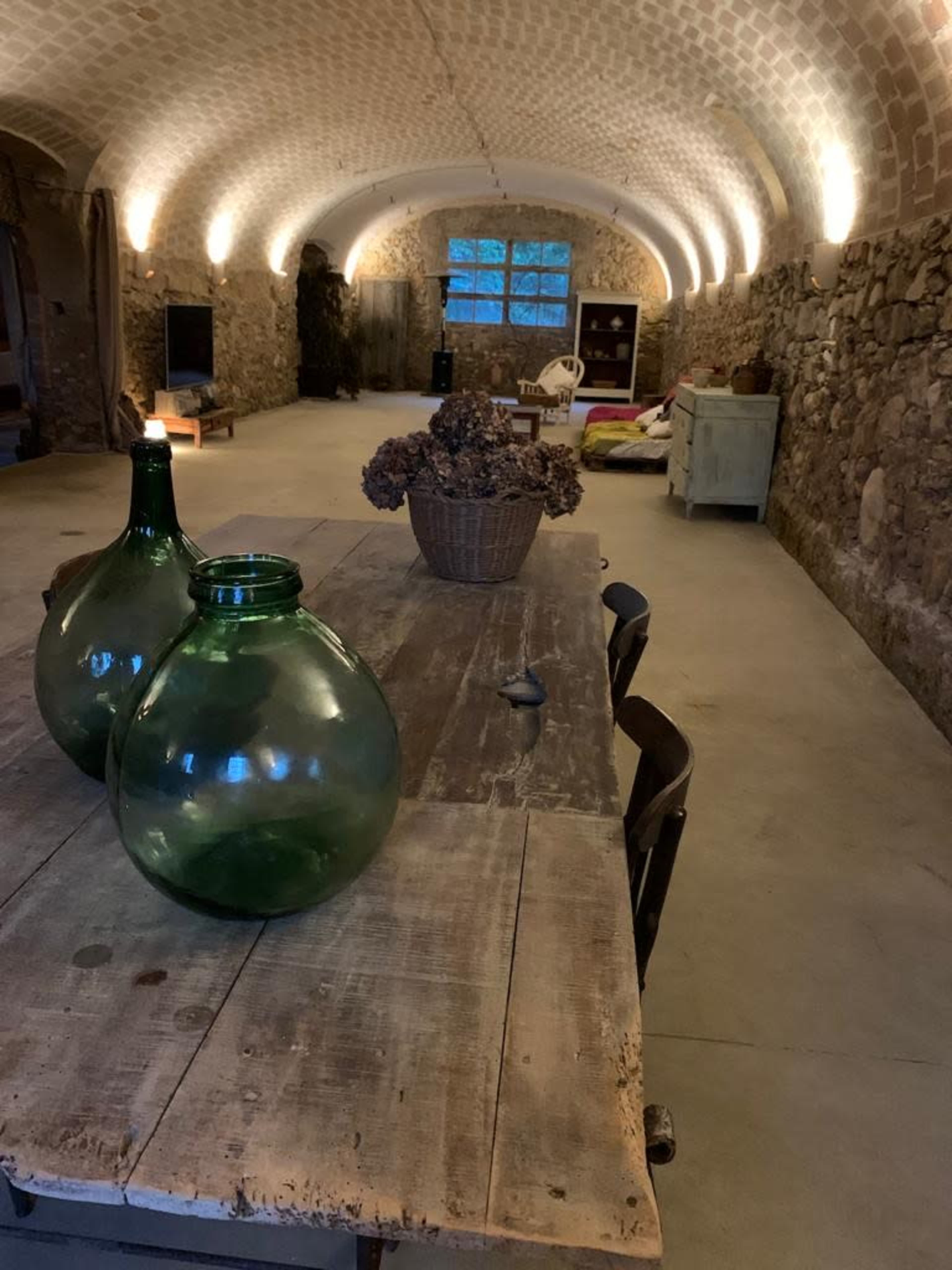 A spacious room with stone walls, a wooden table with green glass bottles, and simple furnishings in a rustic style.