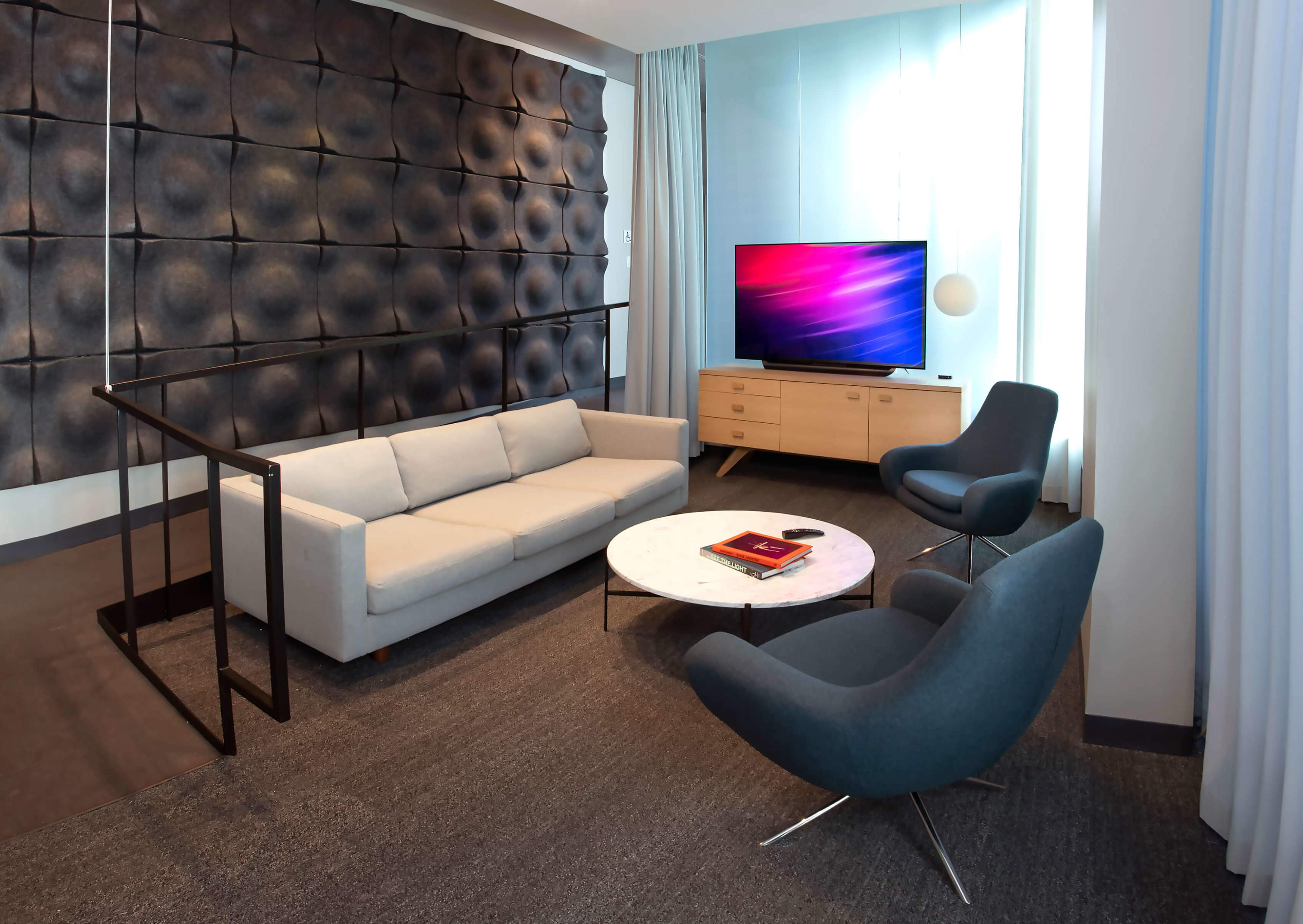 The image shows a modern living room with a gray sofa, two blue chairs, a marble coffee table, and a television against a textured wall.