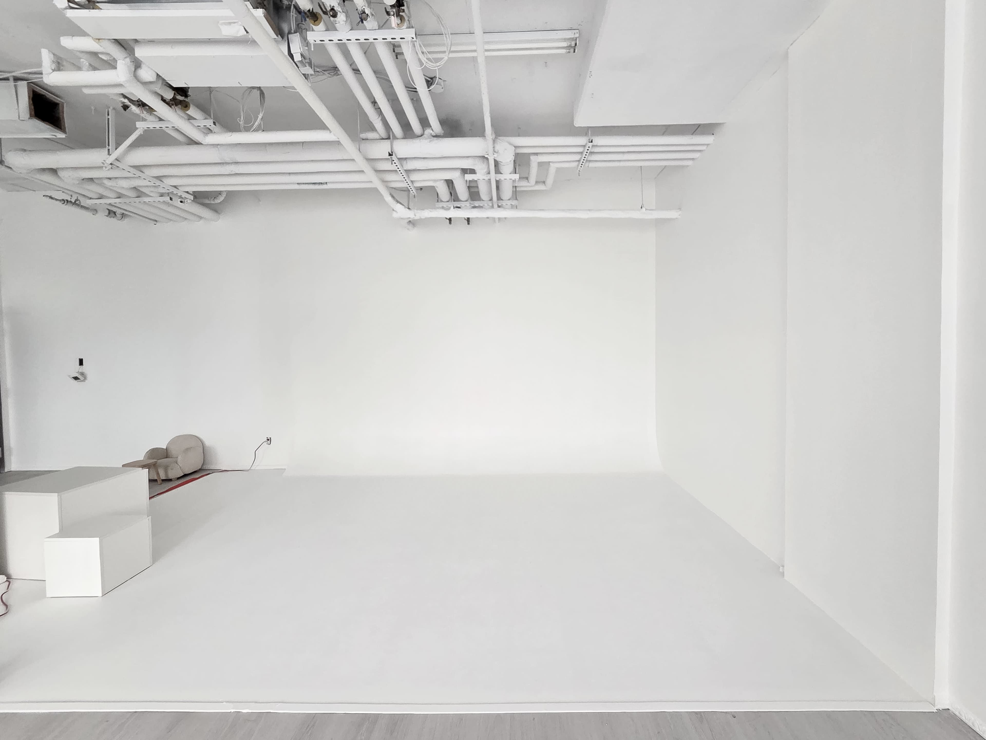The image shows a minimalist, empty room with white walls, a smooth white floor, and visible overhead pipes.