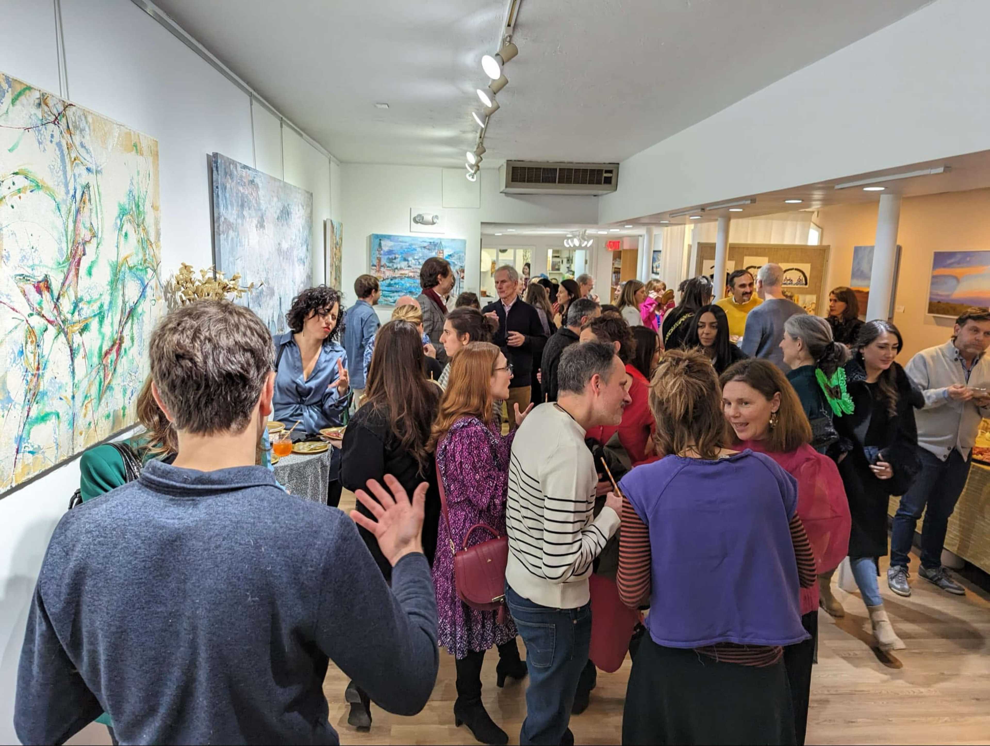 A crowded art gallery opening features people socializing among various displayed artworks.