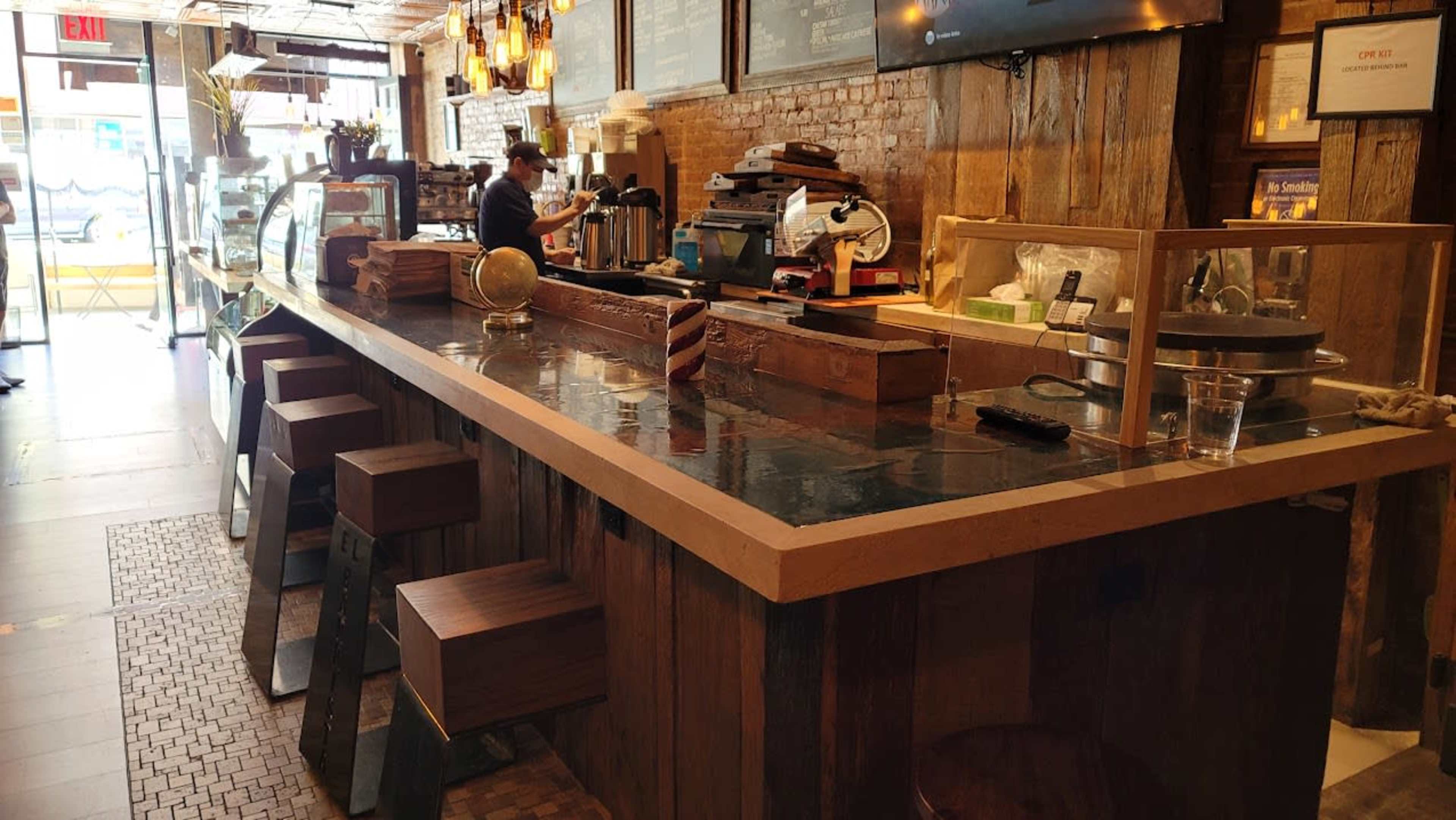 A rustic café interior with a wooden counter, high stools, and a barista preparing drinks behind the counter.