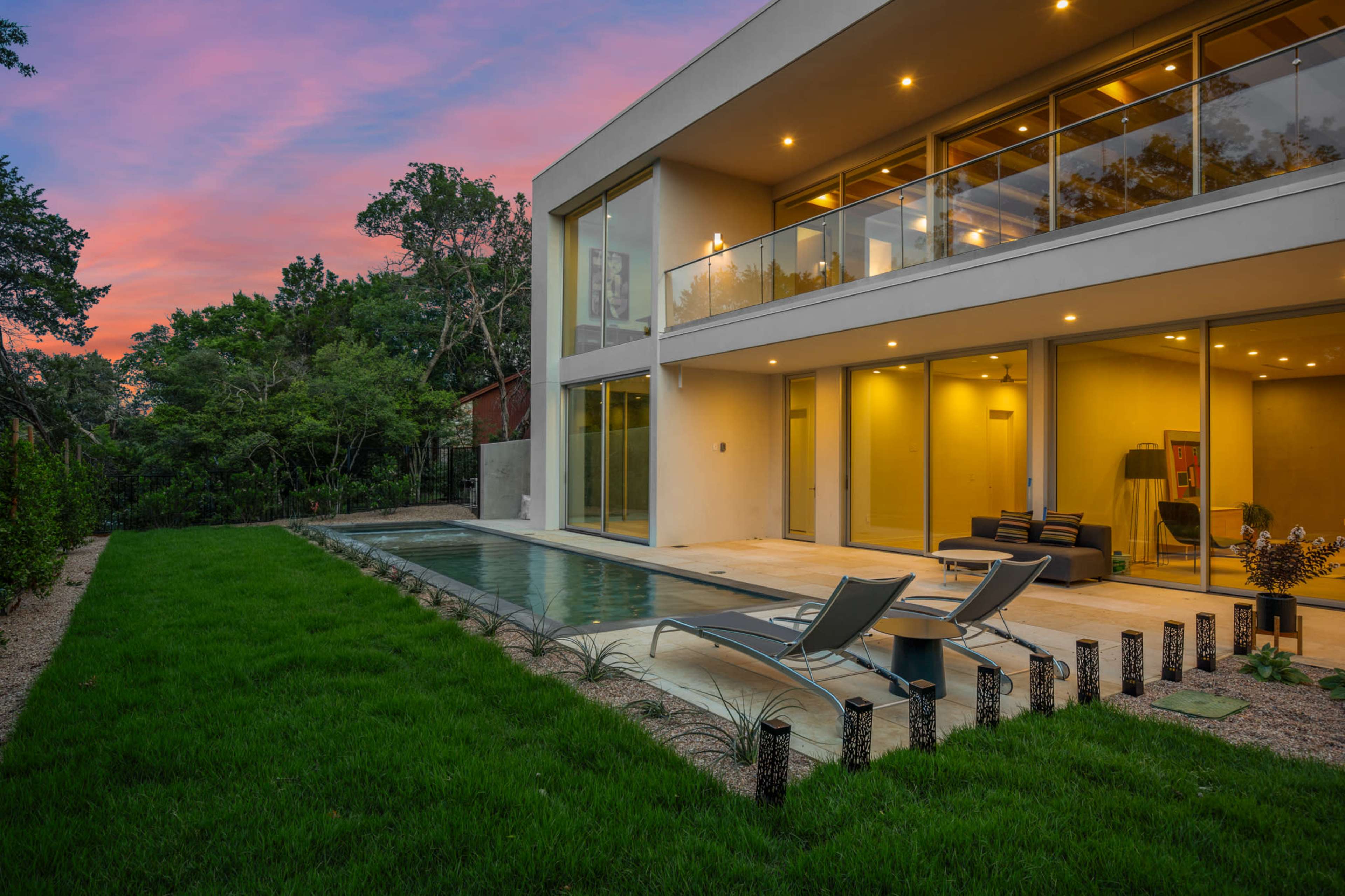 A modern house with large glass windows features a rectangular swimming pool and two lounge chairs on a grassy lawn at sunset.