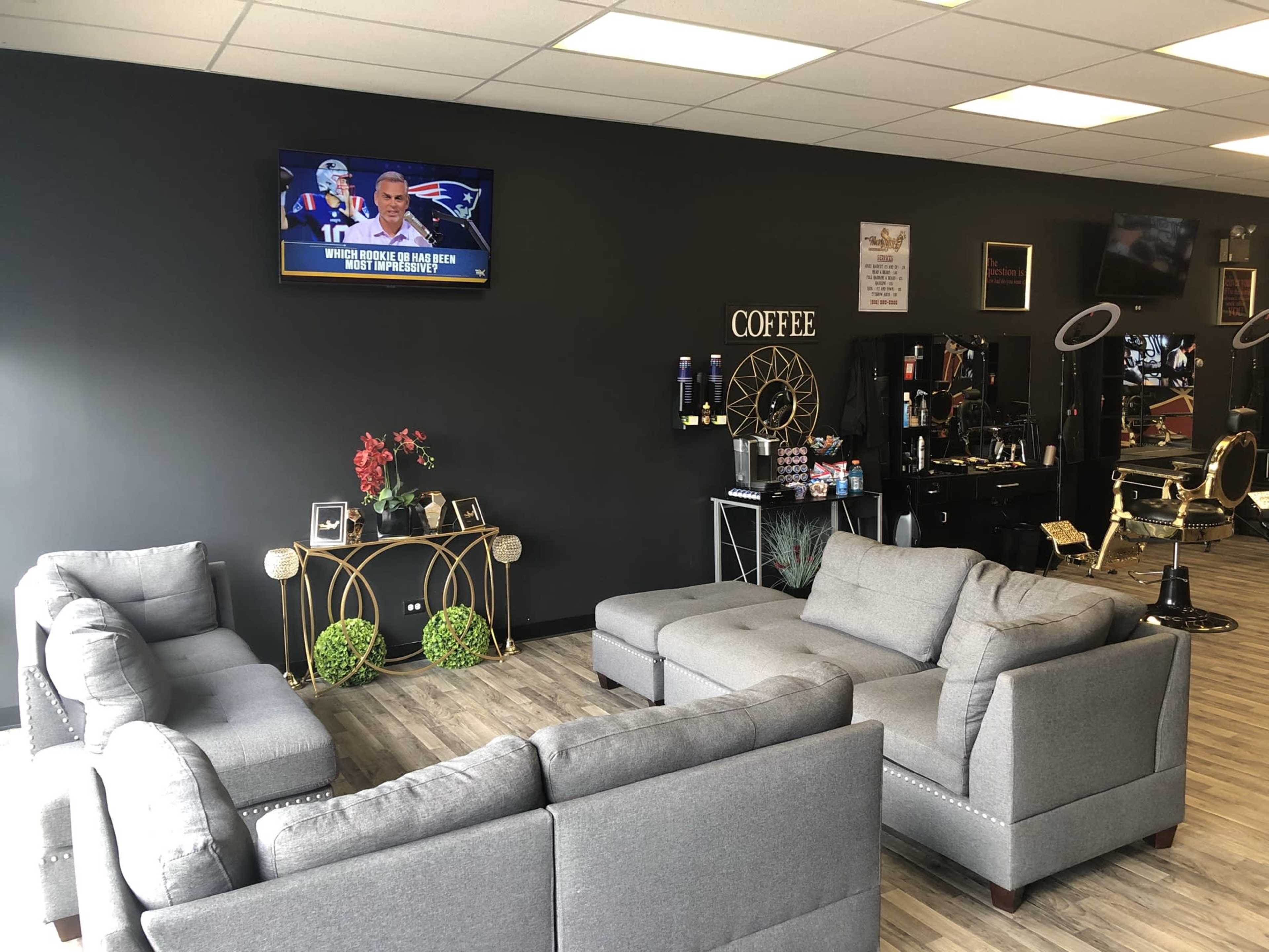 A waiting area with gray sofas, a wall-mounted television displaying a sports program, and a coffee station in a modern barber shop.