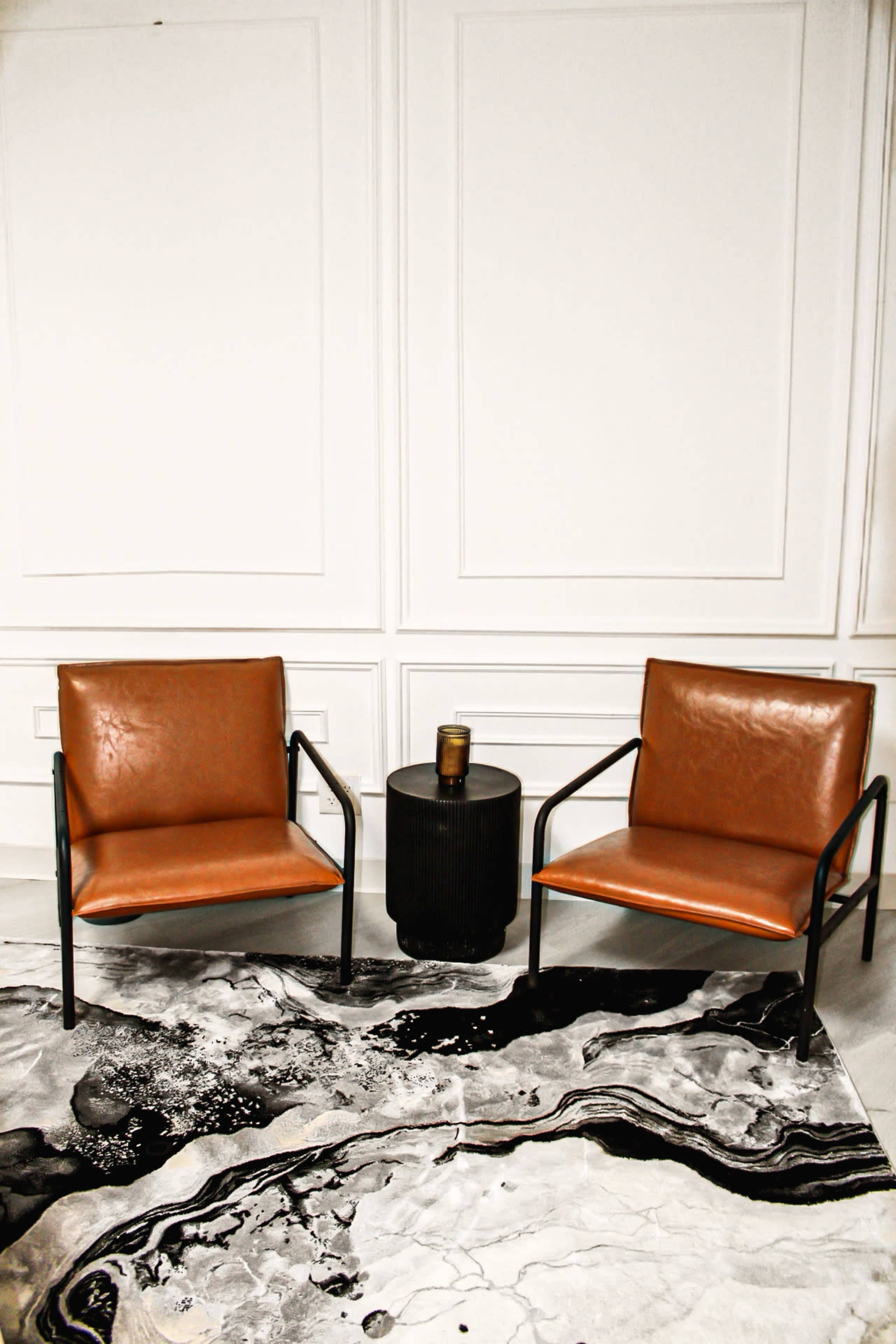 The image features two brown leather chairs flanking a black side table on a patterned rug with abstract designs.